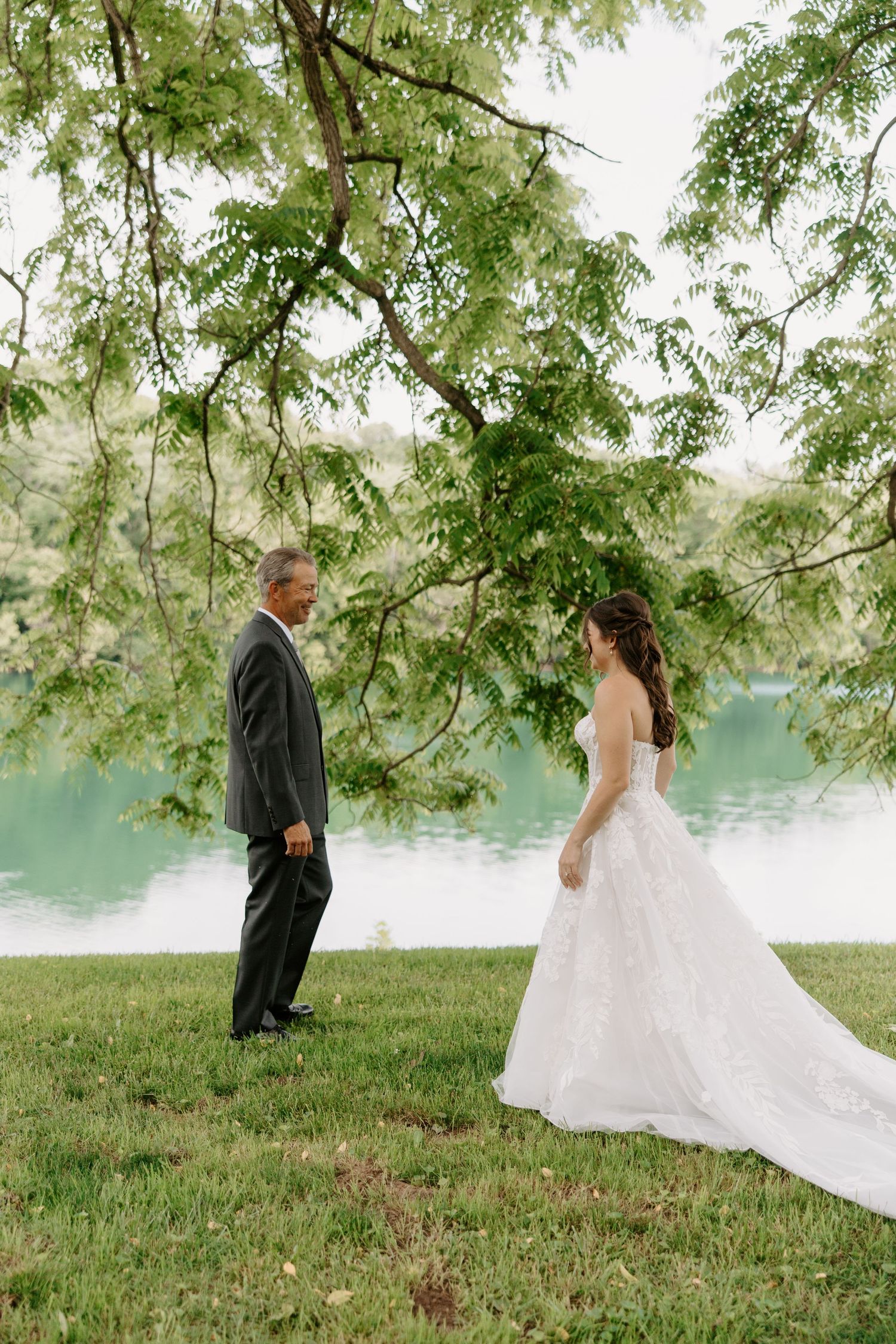 Bride in white lace gown shares first look moment with father by serene lake under lush green tree canopy.