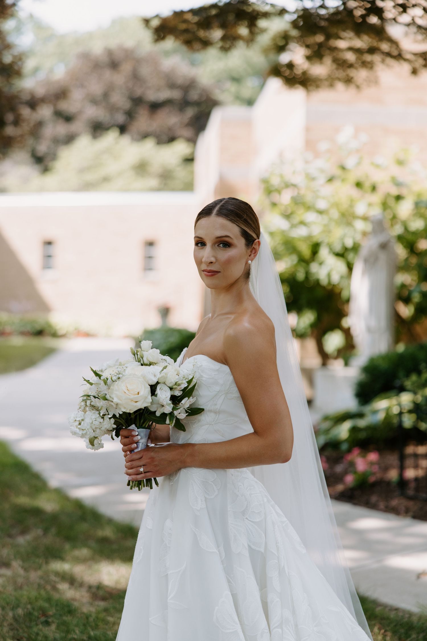 Elegant bride in strapless gown holds white floral bouquet while posing outdoors on wedding day.