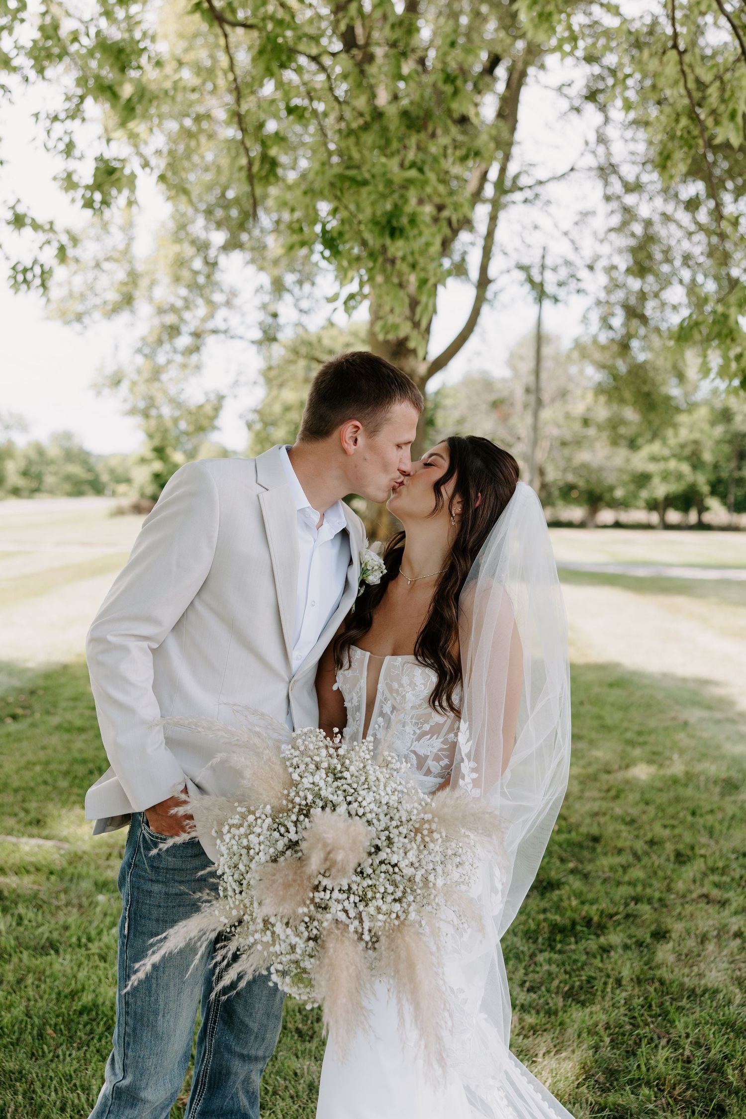Bride and groom share a kiss under a tree, bride holding a rustic pampas grass bouquet.