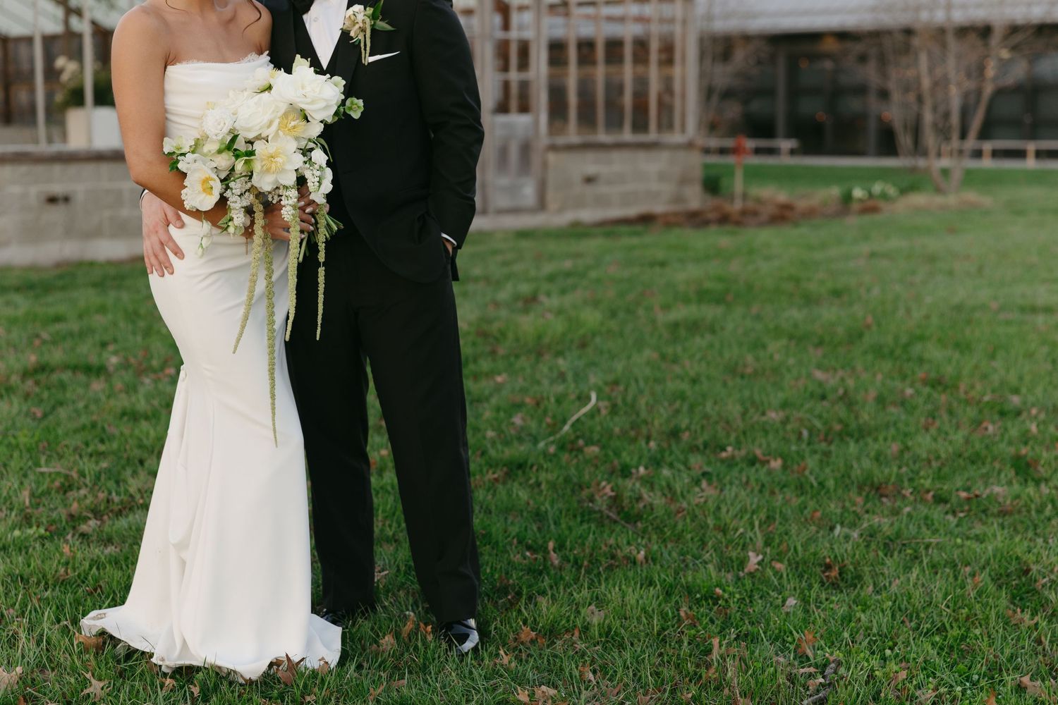 Bride in strapless fitted white gown with white bouquet standing close to groom in black tuxedo on lawn.