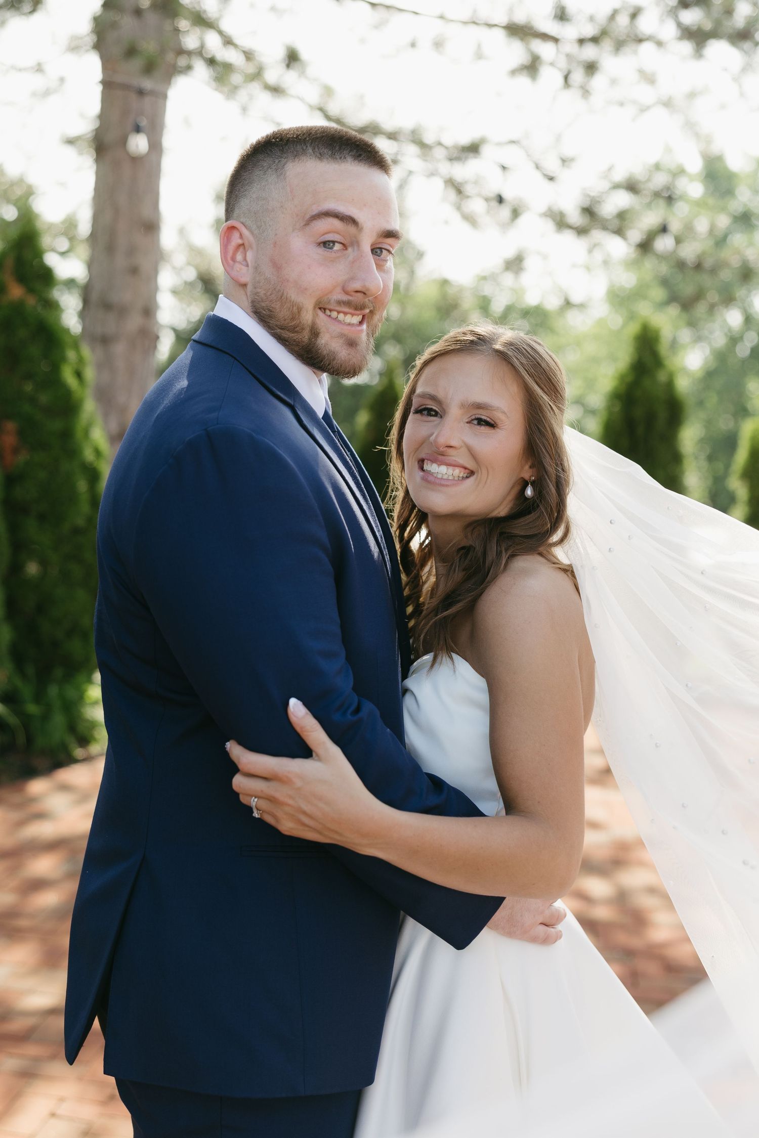 Smiling bride and groom embrace outdoors, groom in navy suit and bride in white gown with flowing veil.