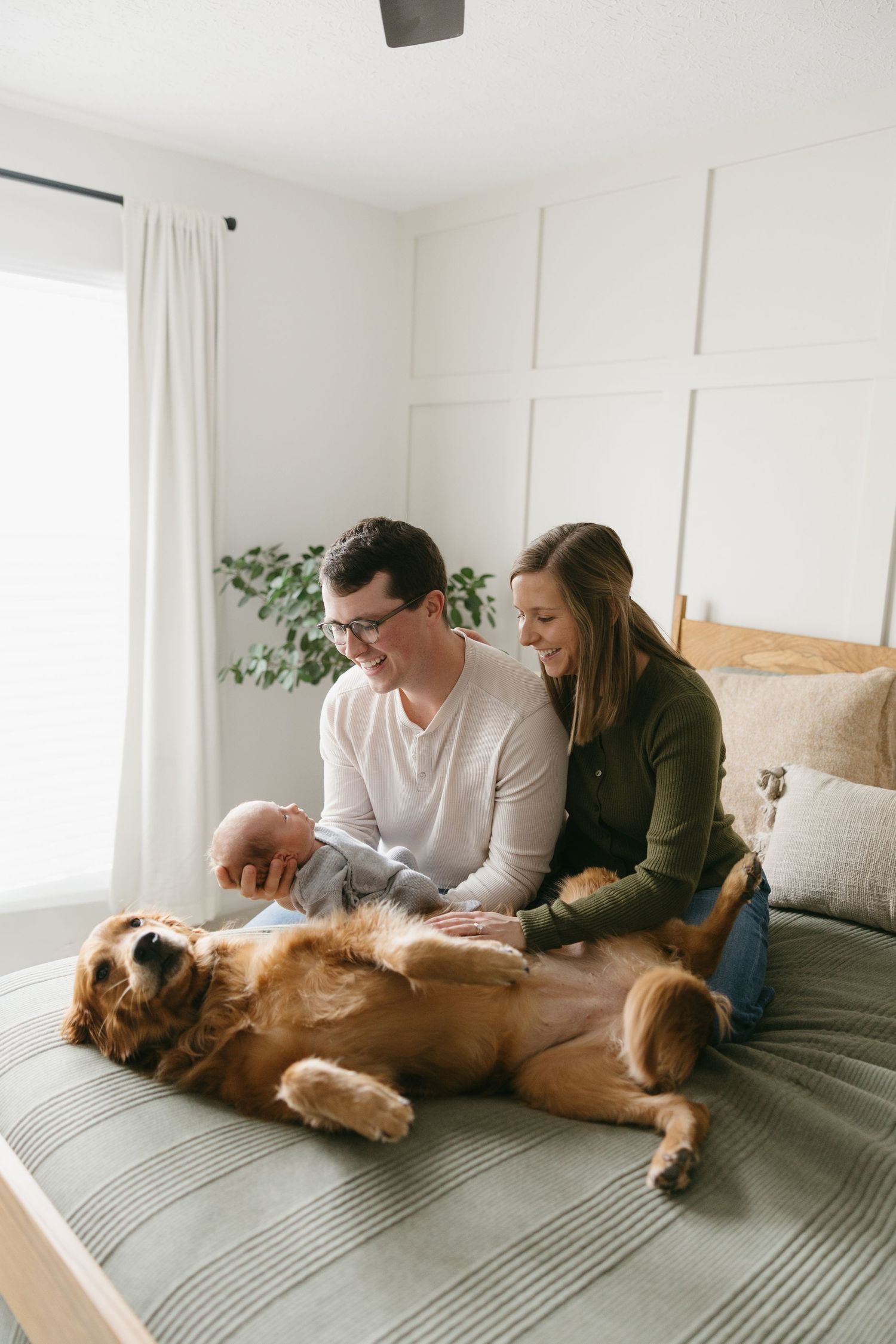 Happy couple sitting on a bed with their golden retriever and newborn baby indoors.