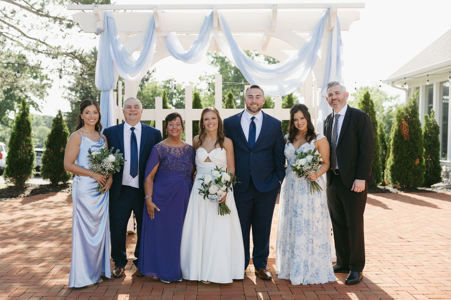 Wedding party poses under blue draped arbor on brick patio with bride in white and guests in blue attire.
