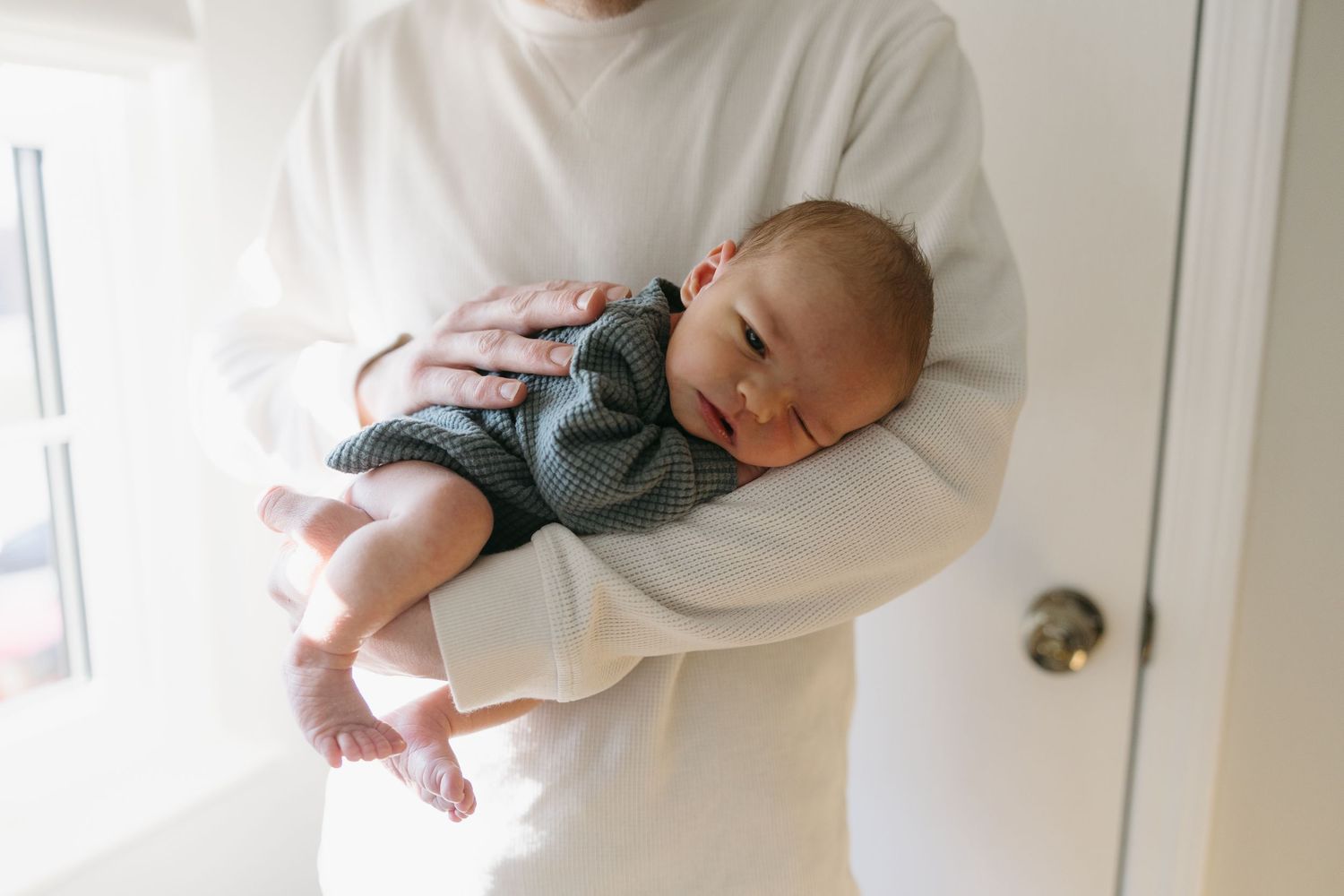 Parent in white sweater gently cradling a sleeping newborn dressed in a gray outfit.