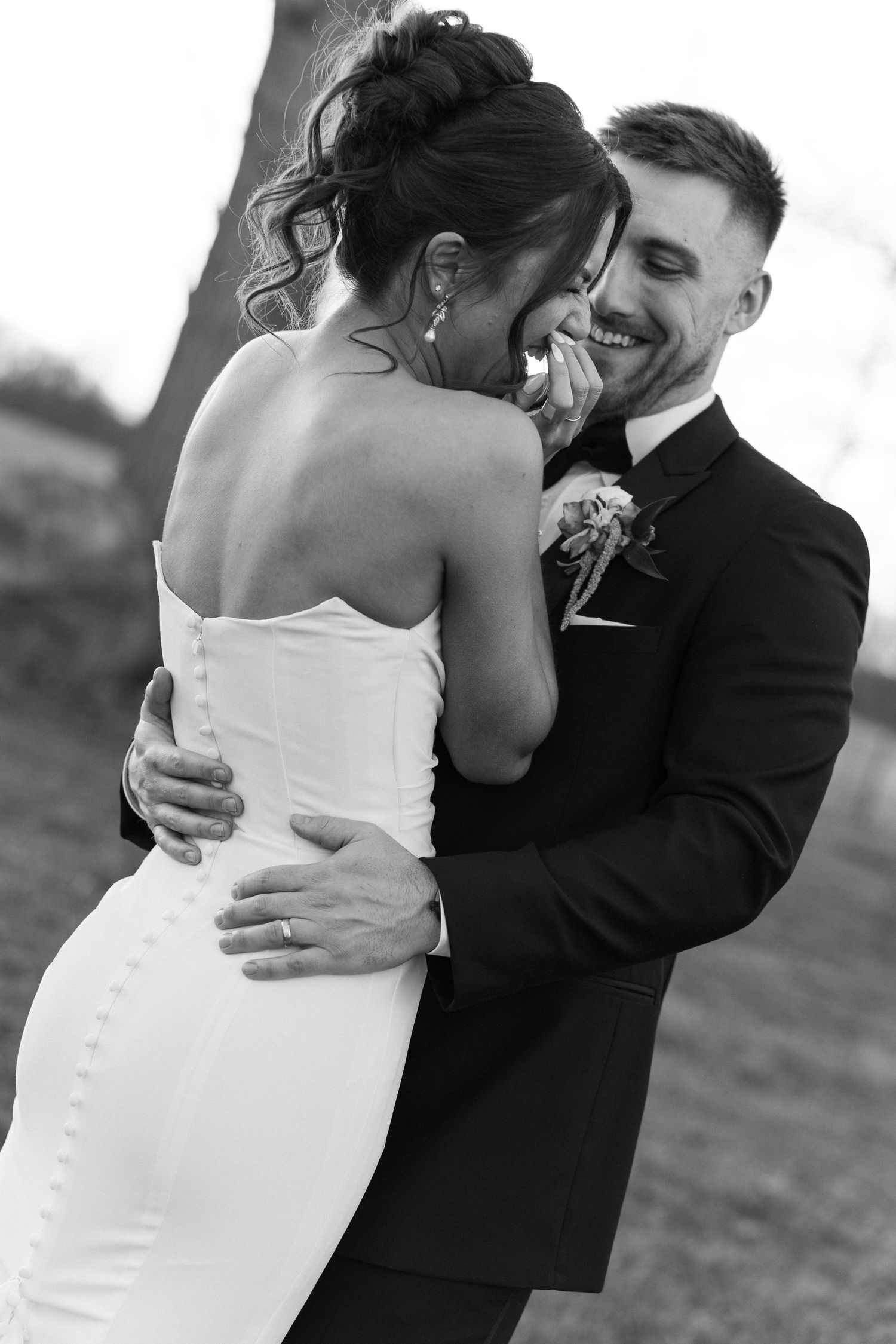 Black and white photo of smiling groom embracing bride in strapless wedding dress outdoors.