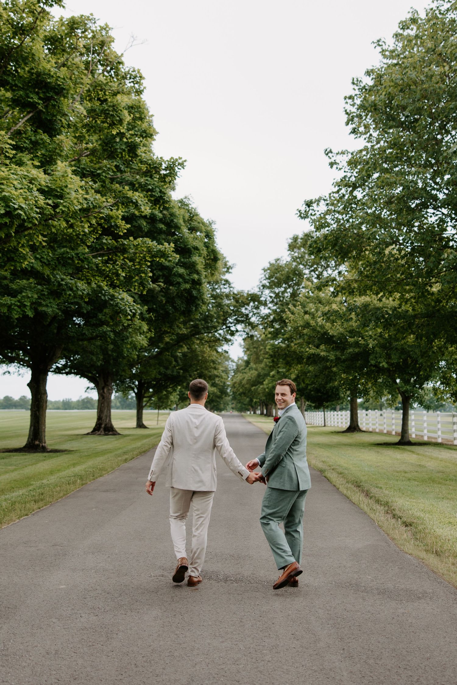 Two grooms in suits hold hands walking down a tree-lined path at an outdoor wedding venue.