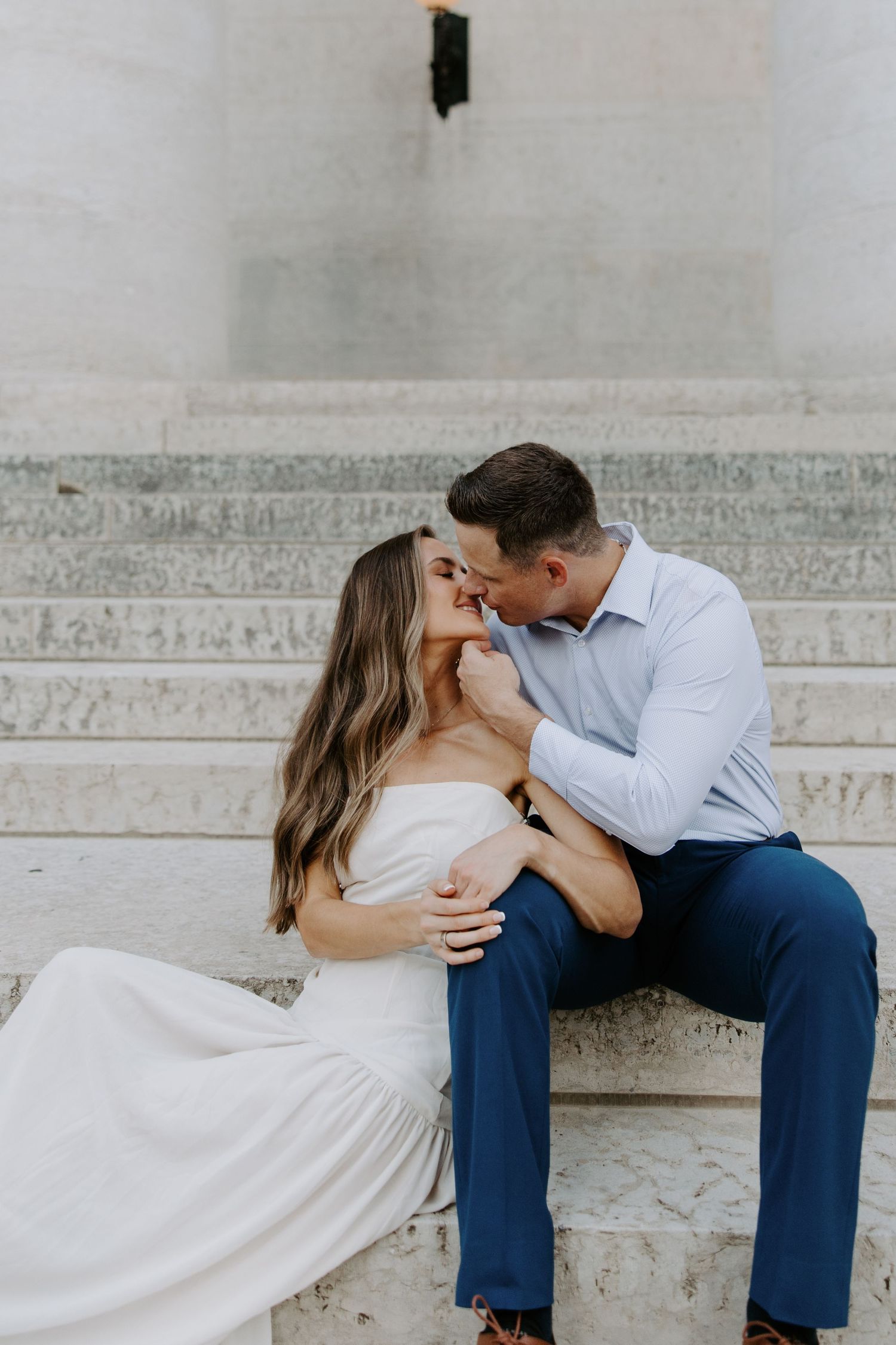 Couple sharing a romantic kiss while seated on marble steps during an outdoor engagement photo session.