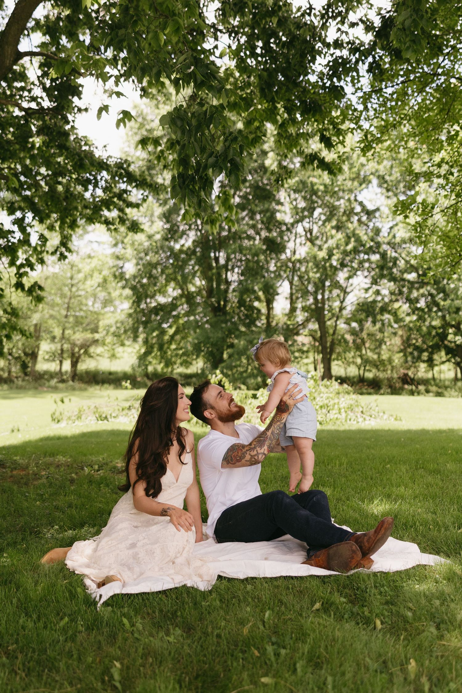 Young tattooed couple sits on blanket in park, lifting smiling baby boy under leafy green trees.