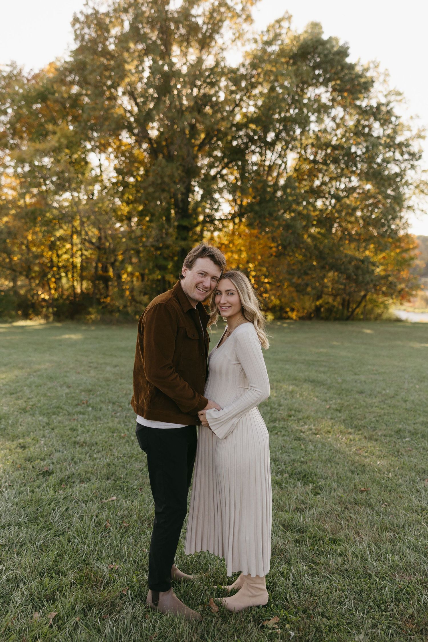 Couple embracing in a green field surrounded by autumn trees during golden hour in a fall engagement photo.