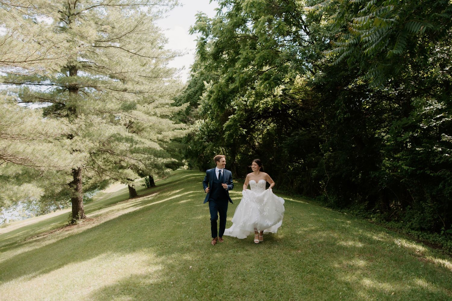 Bride and groom walking hand in hand across lush green lawn surrounded by tall trees on their wedding day.