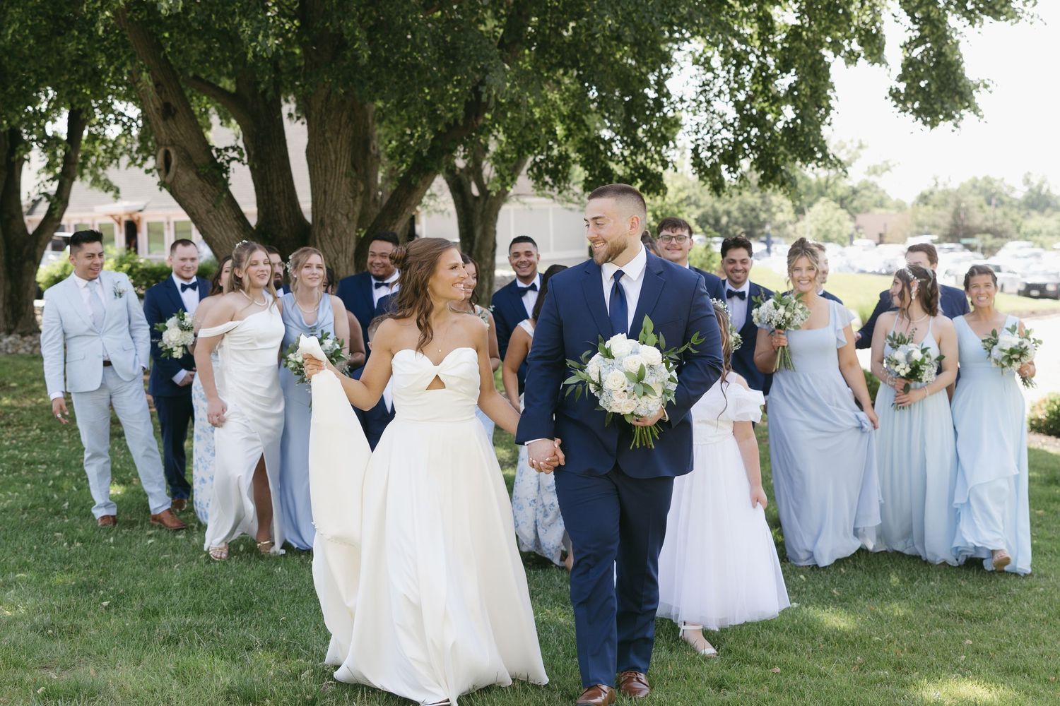 Bride and groom walk with their wedding party in blue and gray attire under large oak trees.