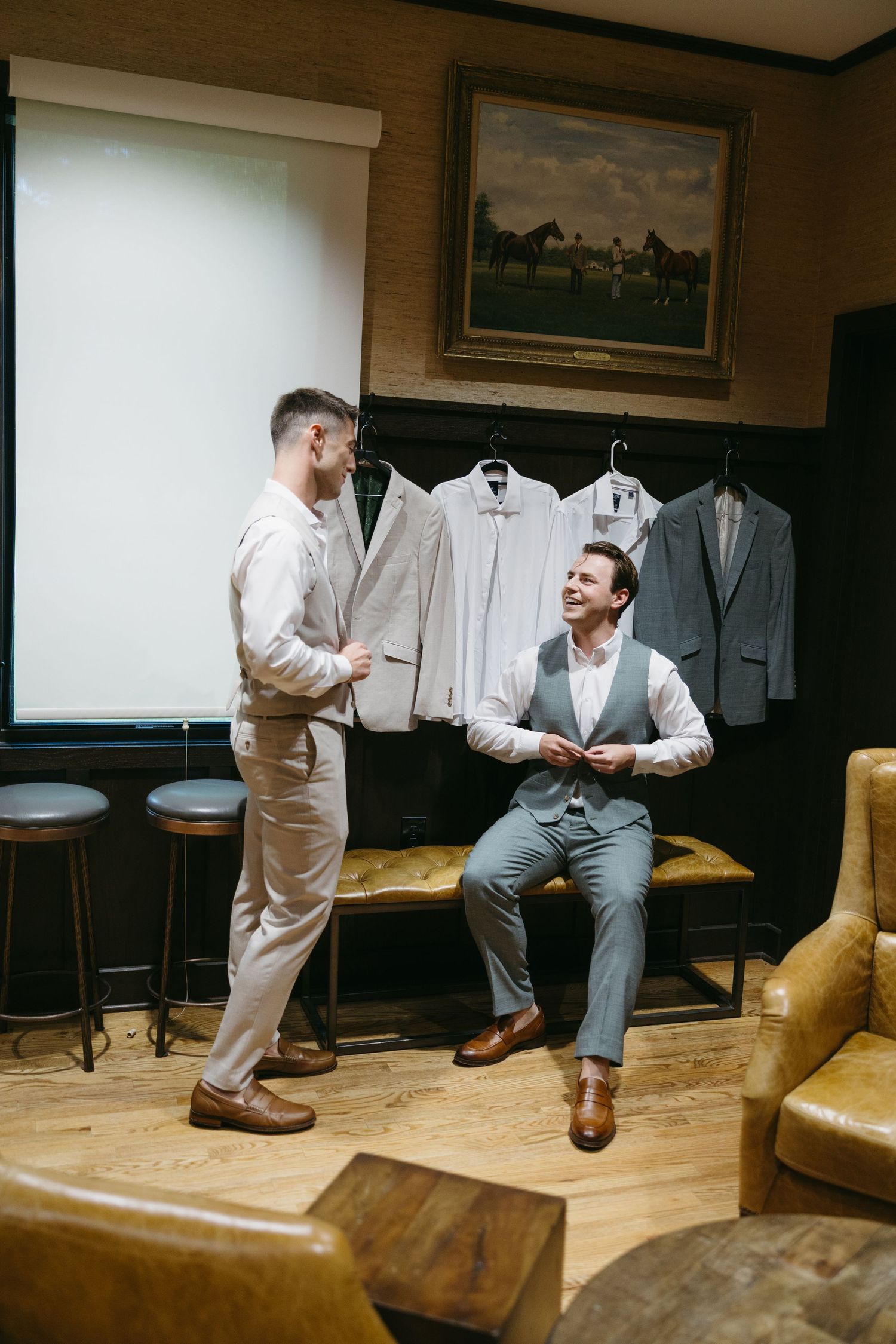 Two well-dressed men in suits chat in an elegant room with hanging garments and leather furniture.