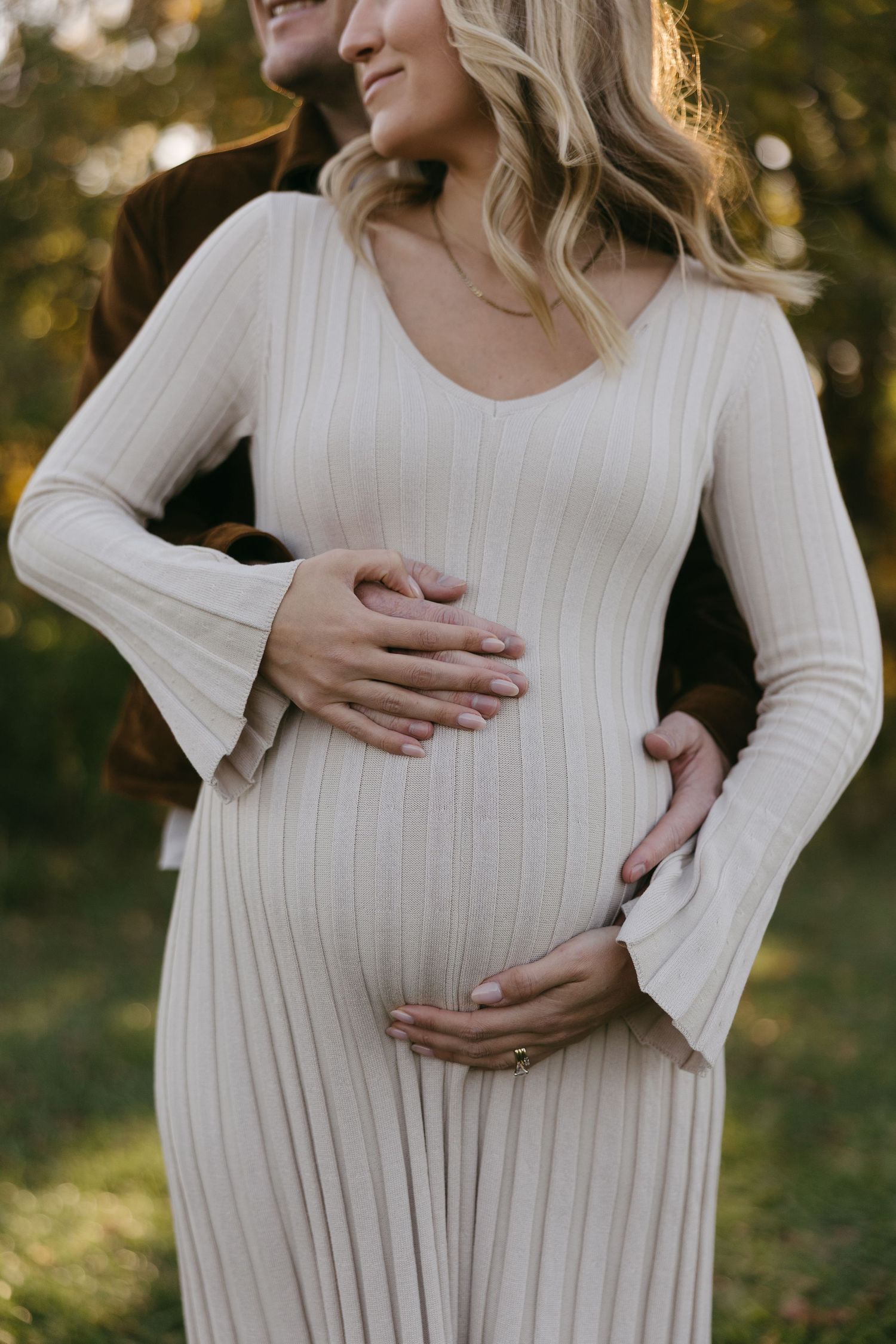 Pregnant woman in ribbed cream dress with partner's hands cradling baby bump during maternity photo shoot outdoors.
