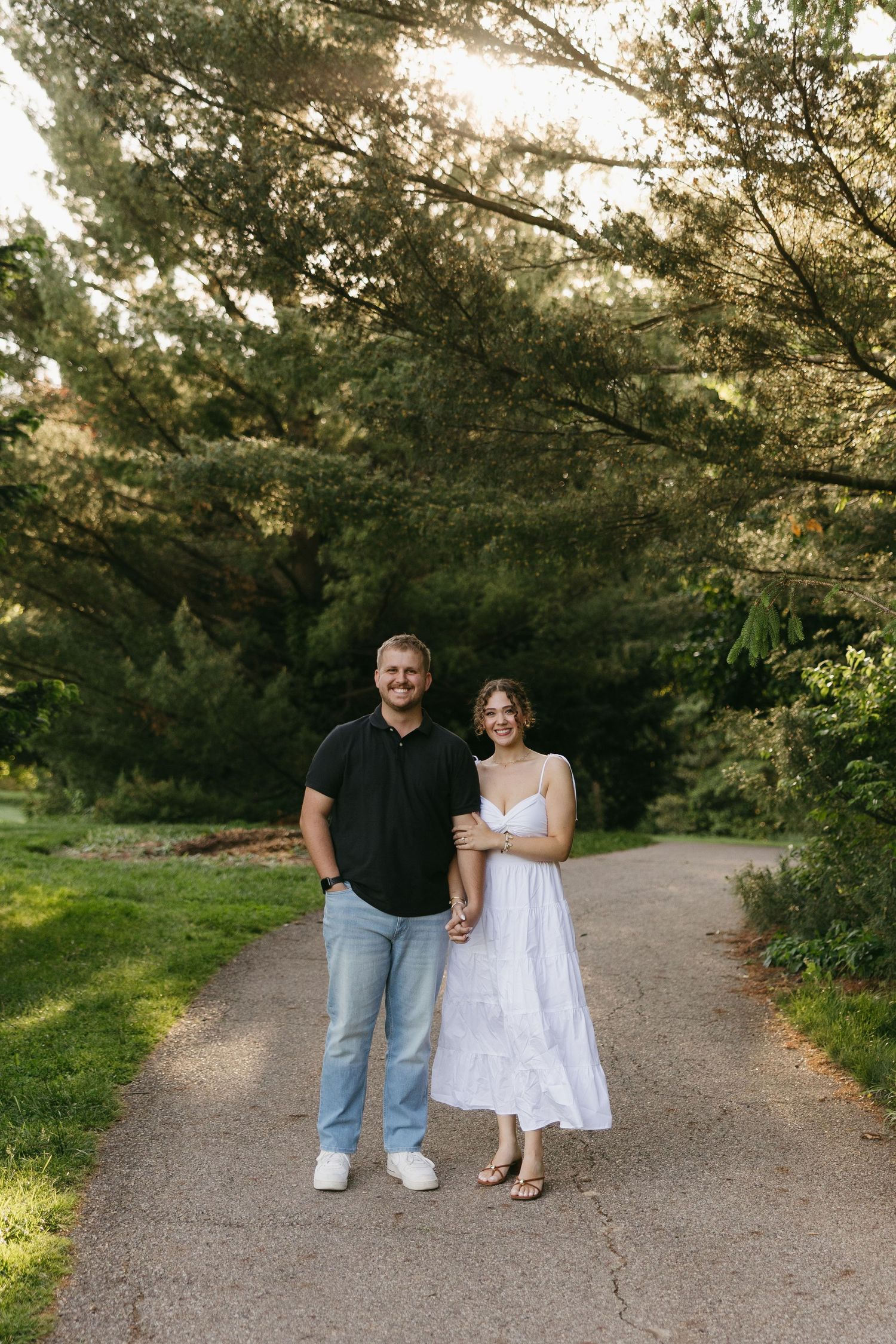 A smiling couple holds hands on a tree-lined path, man in black polo and jeans, woman in white sundress.