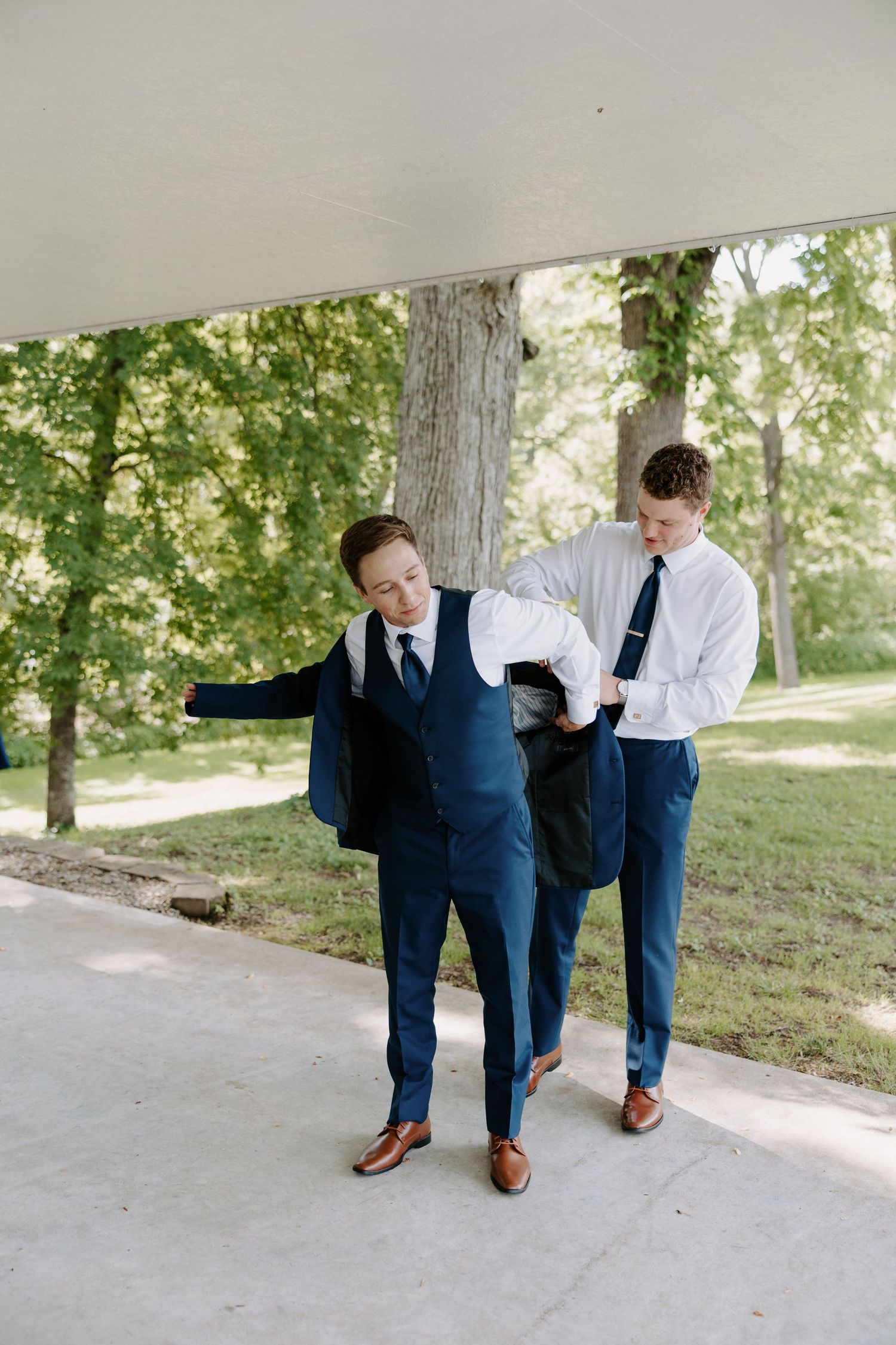 Two groomsmen in navy blue suits and black ties prepare for a wedding ceremony outdoors near lush green trees.