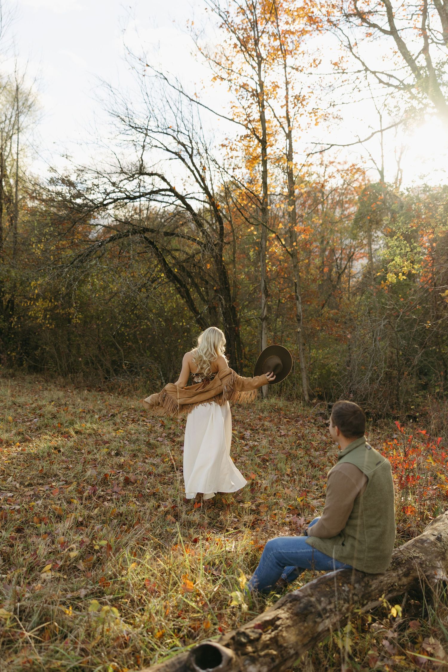 Couple in autumn forest, woman in white dress holding hat, man sitting on log, fall foliage background.