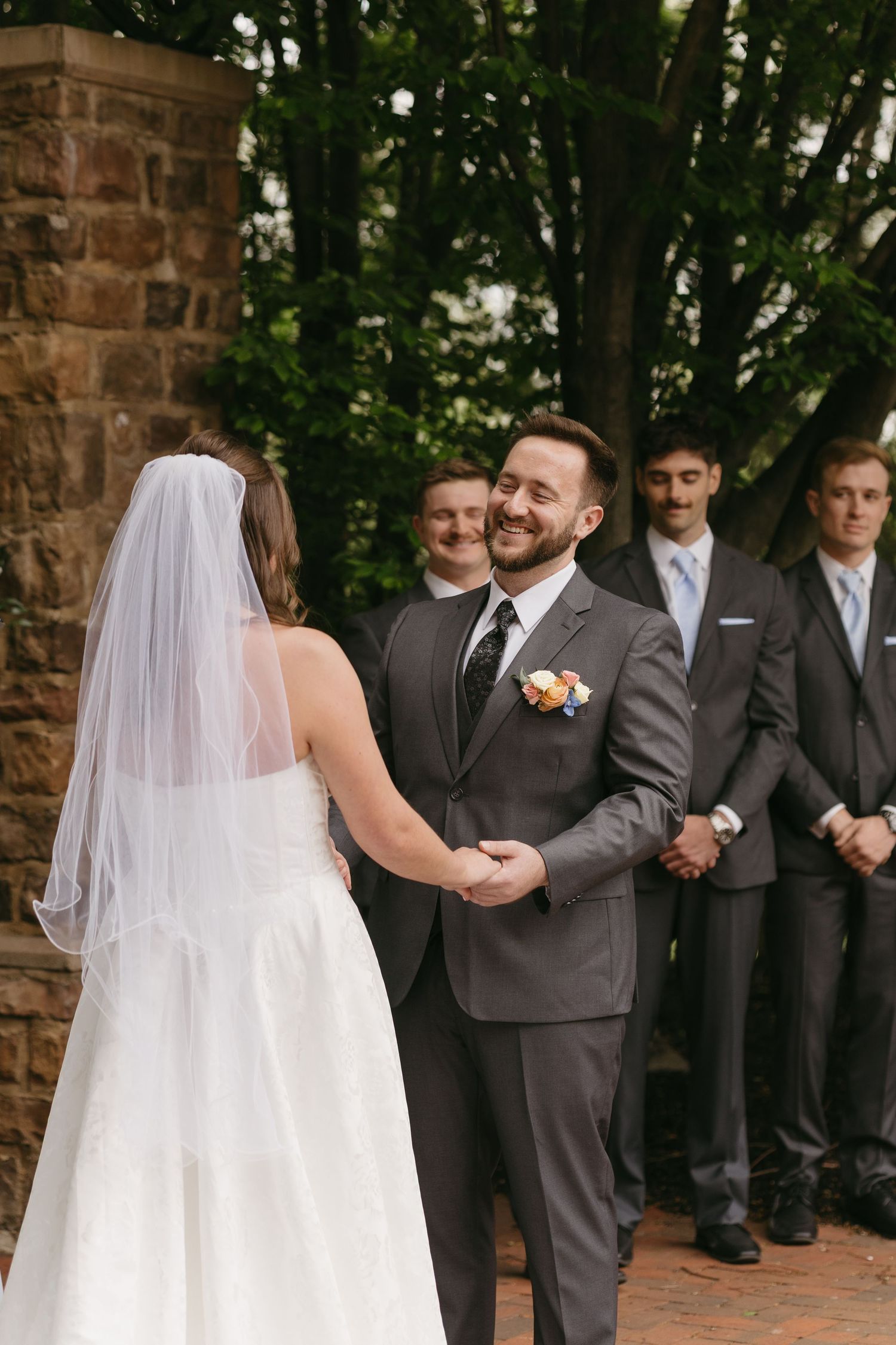Groom smiling warmly at bride during outdoor wedding ceremony with groomsmen standing behind him.