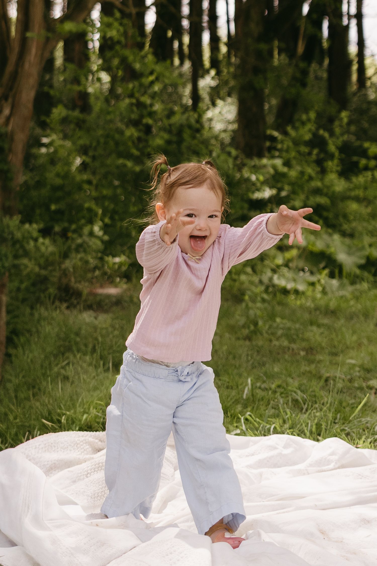 Toddler girl in pink top and jeans laughing and reaching out on white blanket in green park.