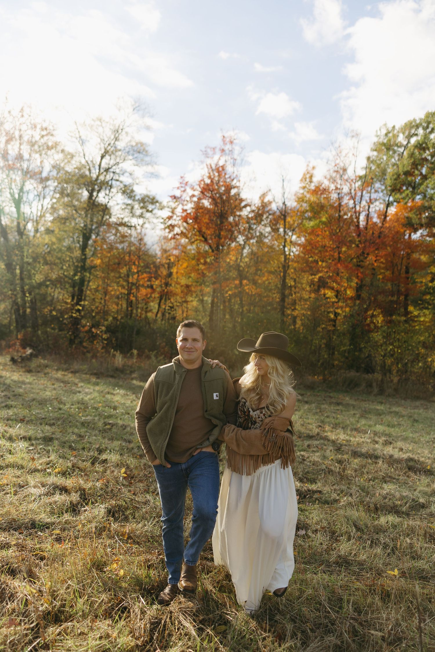 Couple walking in autumn field, woman in white dress and hat, man in green jacket, colorful fall foliage background.