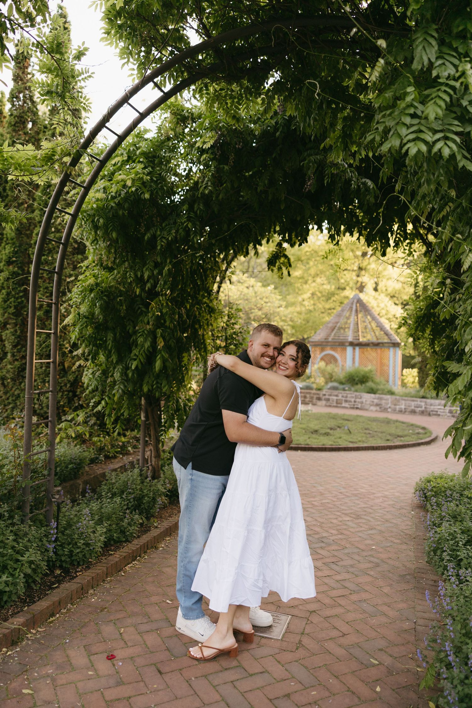 Couple embracing under a green garden arch on a brick path, woman in white dress, man in black shirt.