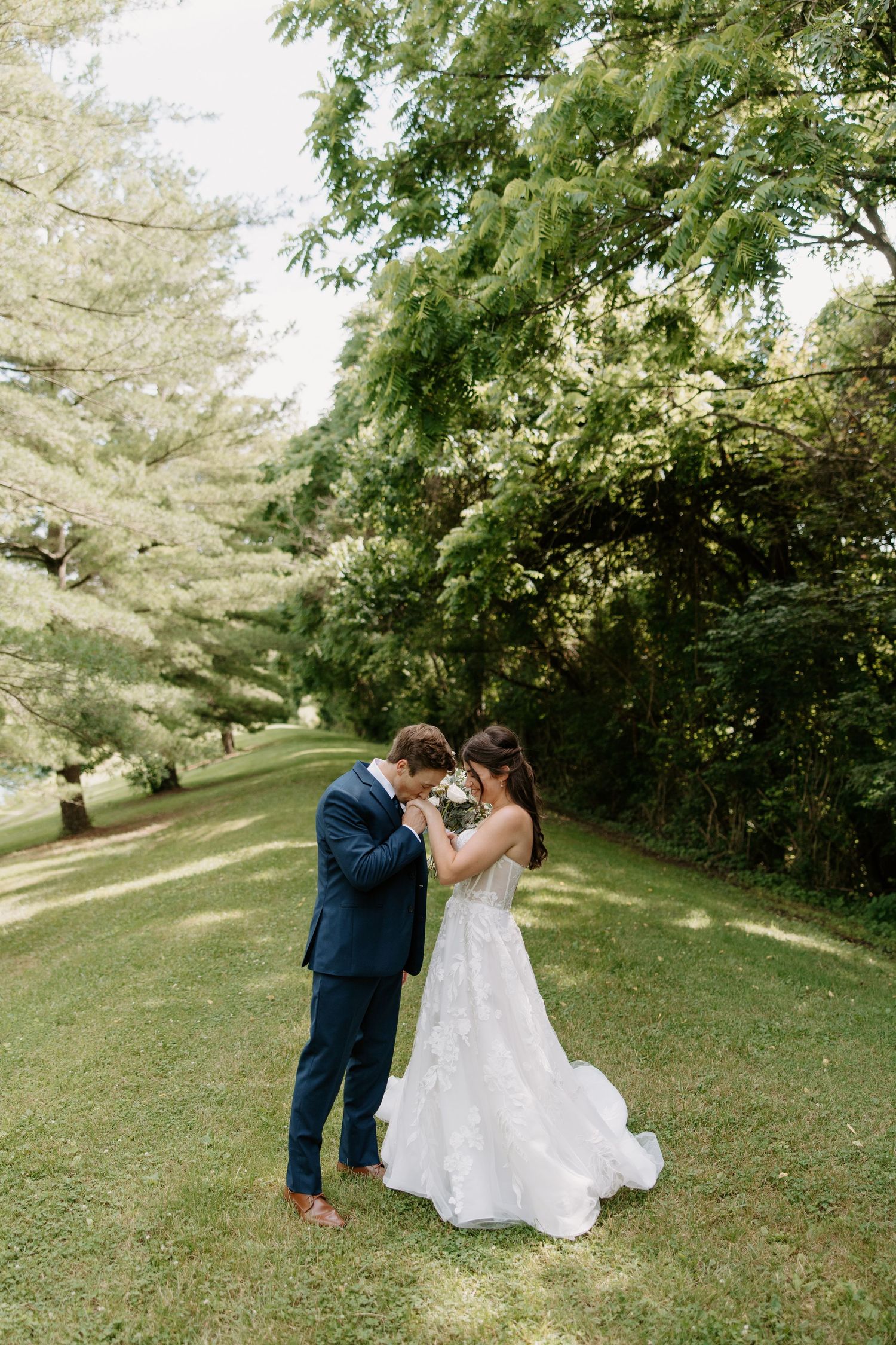 A bride and groom share an intimate moment on a green lawn surrounded by lush trees on their wedding day.