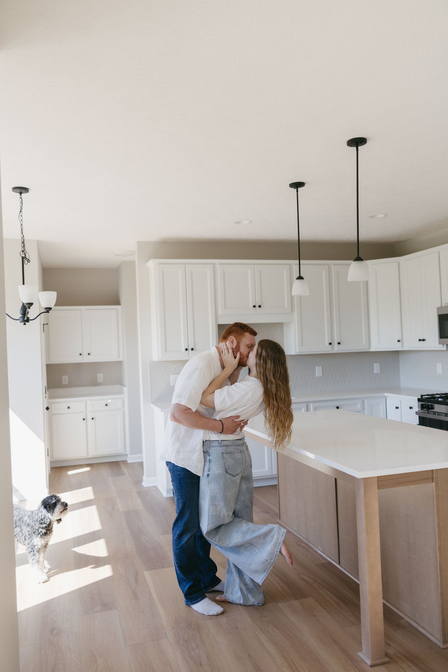 Couple sharing a kiss in a bright modern kitchen with white cabinets and a dog nearby.