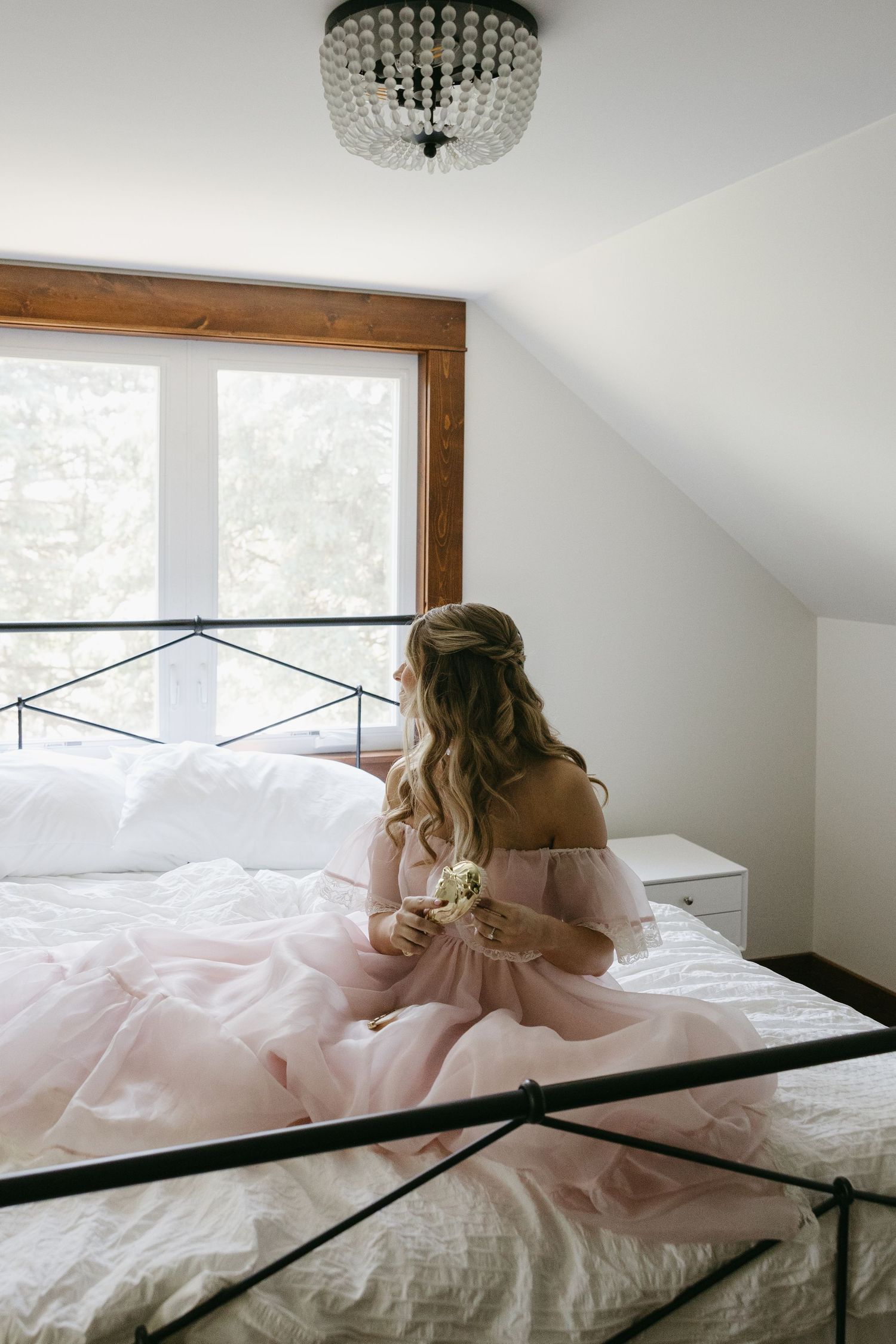 Woman in pink tulle dress sitting on white bed, holding flowers, looking out wooden-framed window in elegant bedroom.
