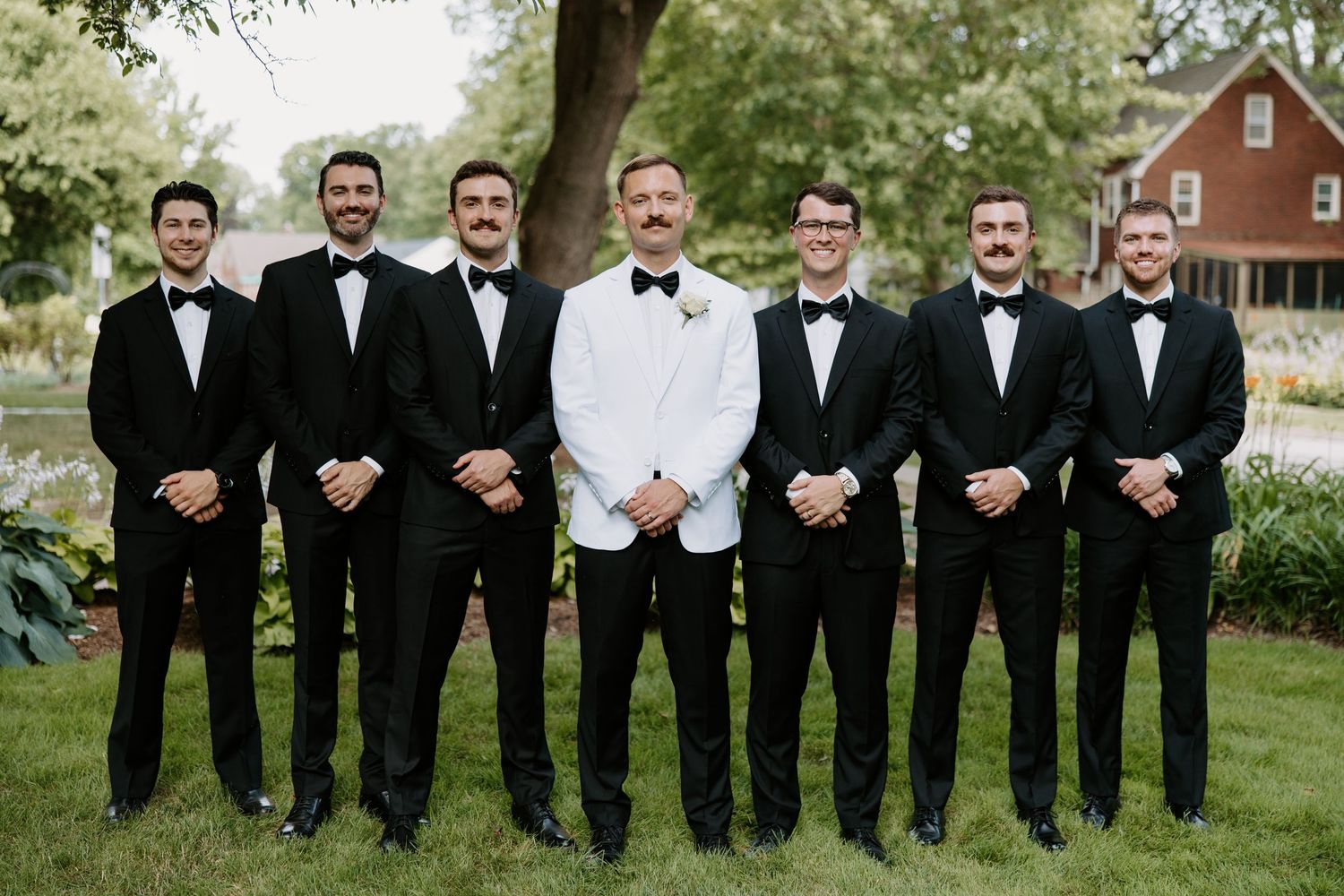 Groom in white tuxedo jacket poses with six groomsmen in black tuxedos outdoors on wedding day.