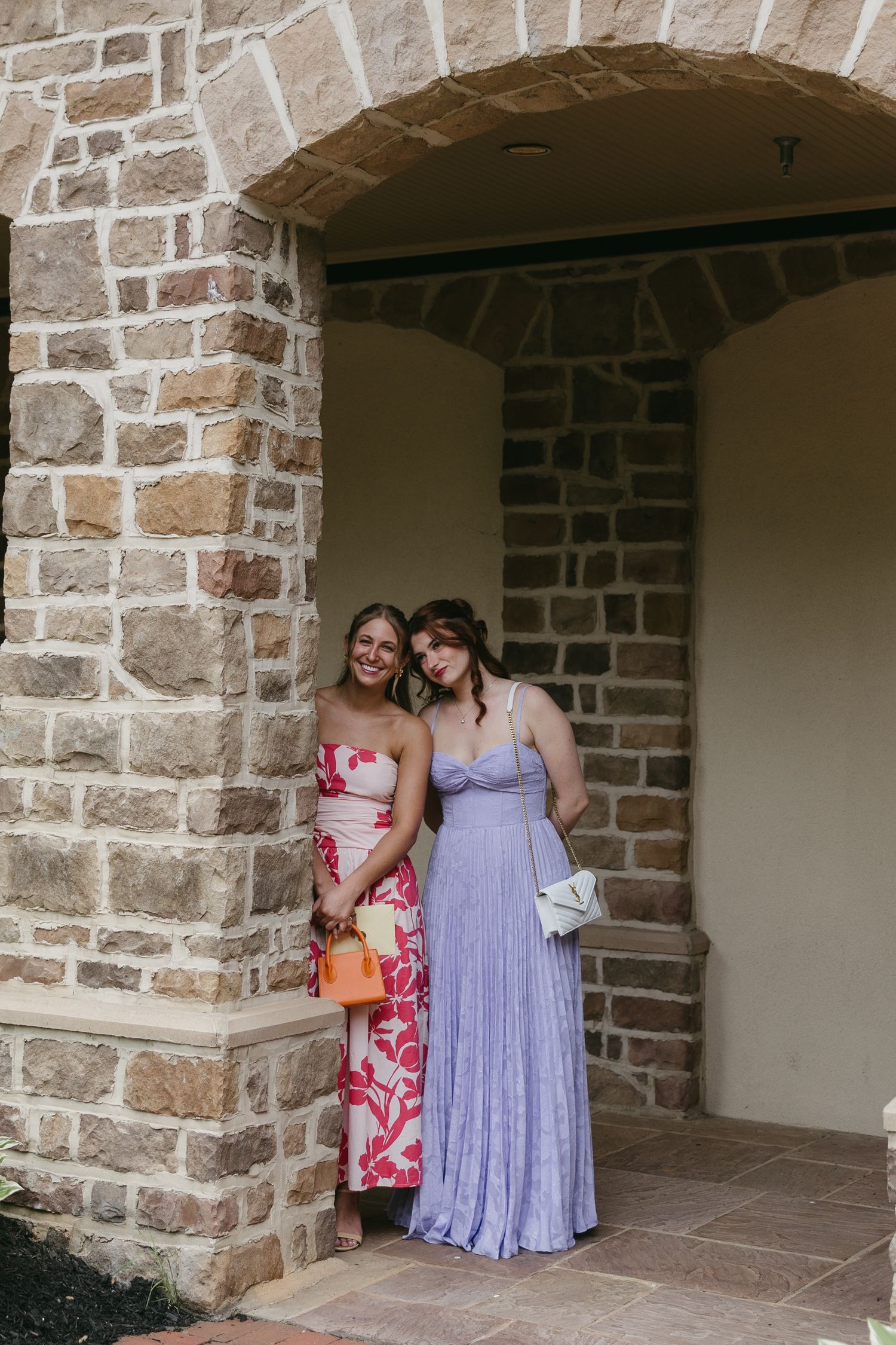 Two female wedding guests posing together in elegant dresses under a stone archway at a wedding venue.