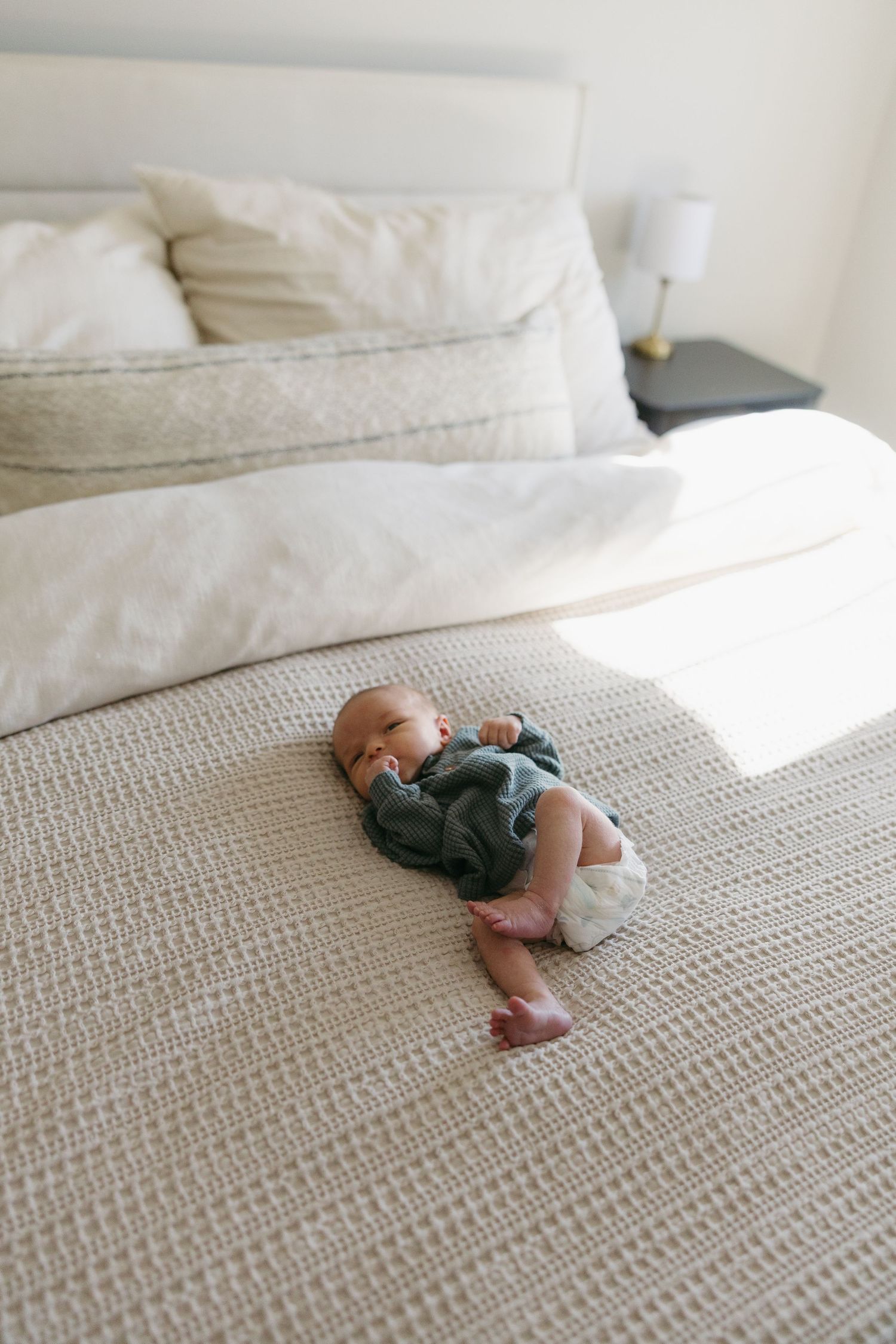 Newborn baby in green onesie lying on a neutral textured bedspread in a bright, minimalist bedroom.