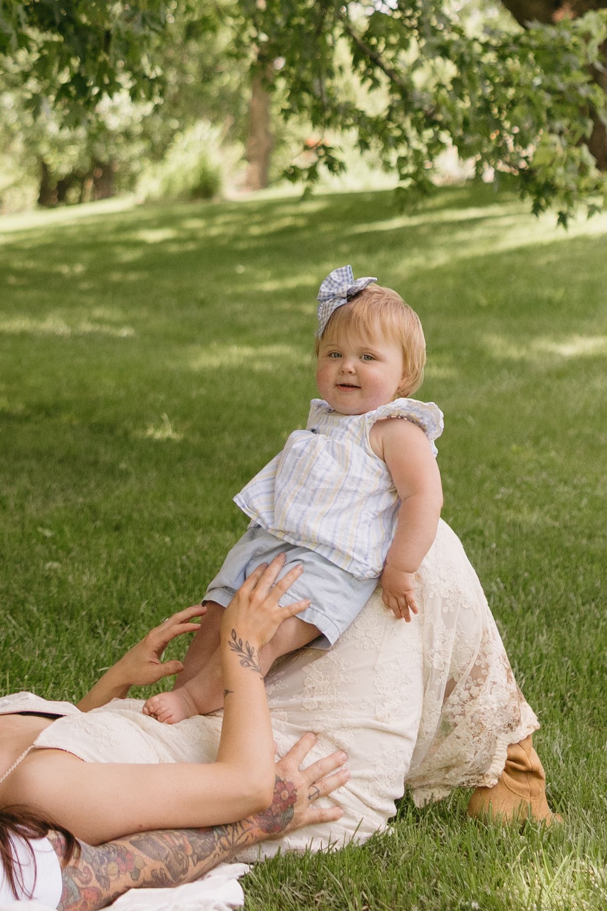 Cheerful baby girl in blue outfit sits on mothers lap on grass blanket in sunny green park.