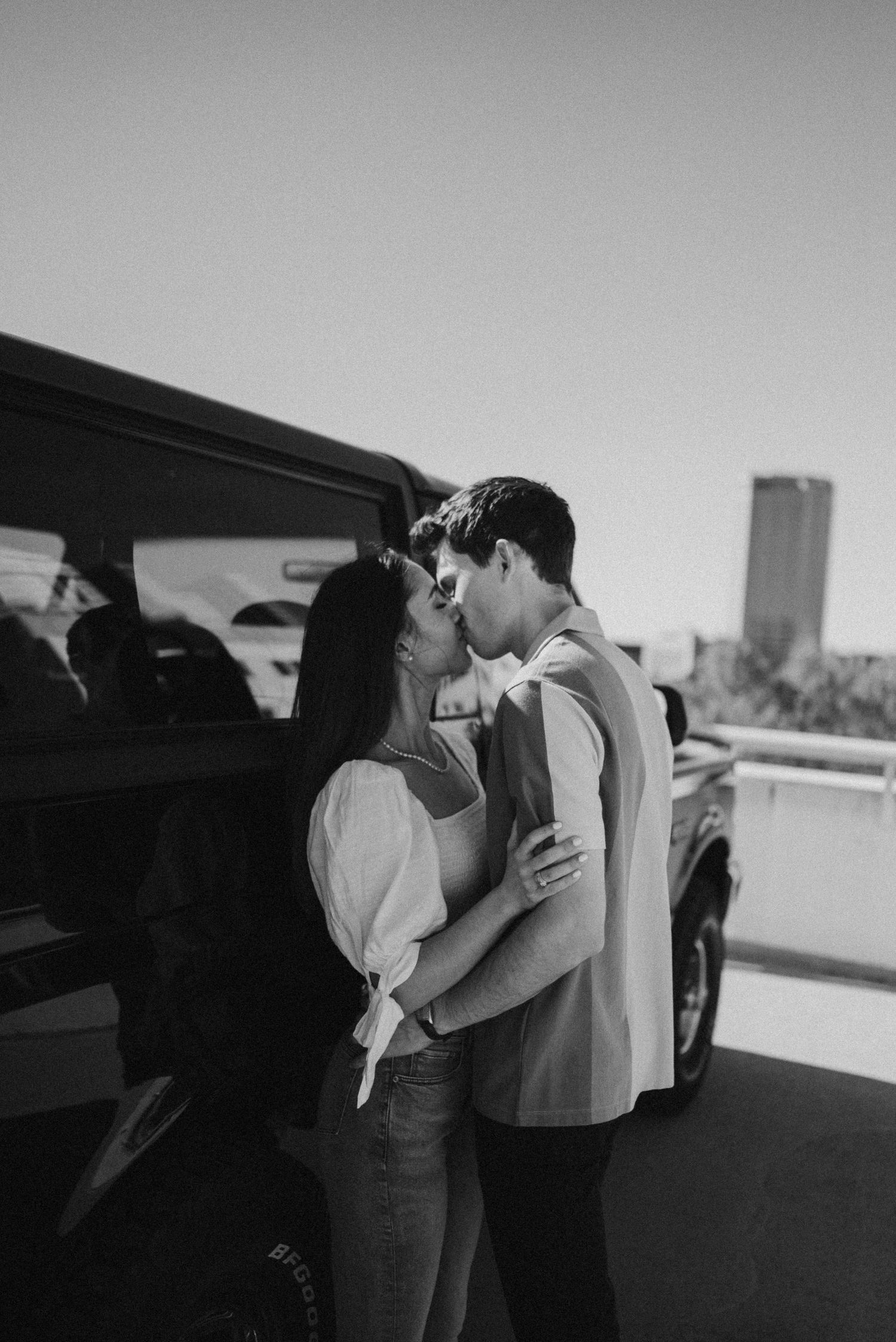 Black and white photo of a couple sharing a kiss beside a dark SUV in an urban setting.