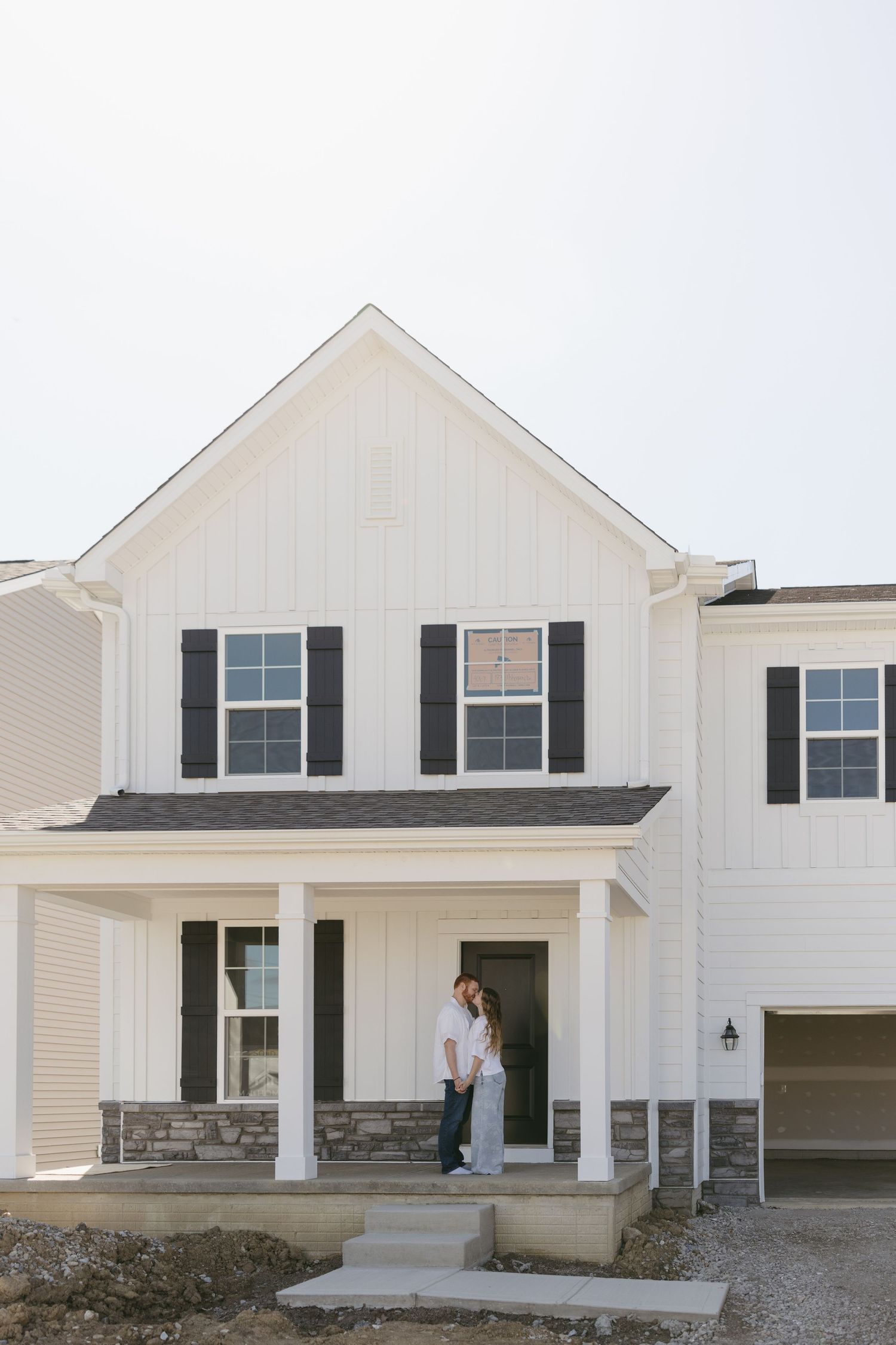 Couple embracing on the front porch of a new white farmhouse-style home with black shutters.
