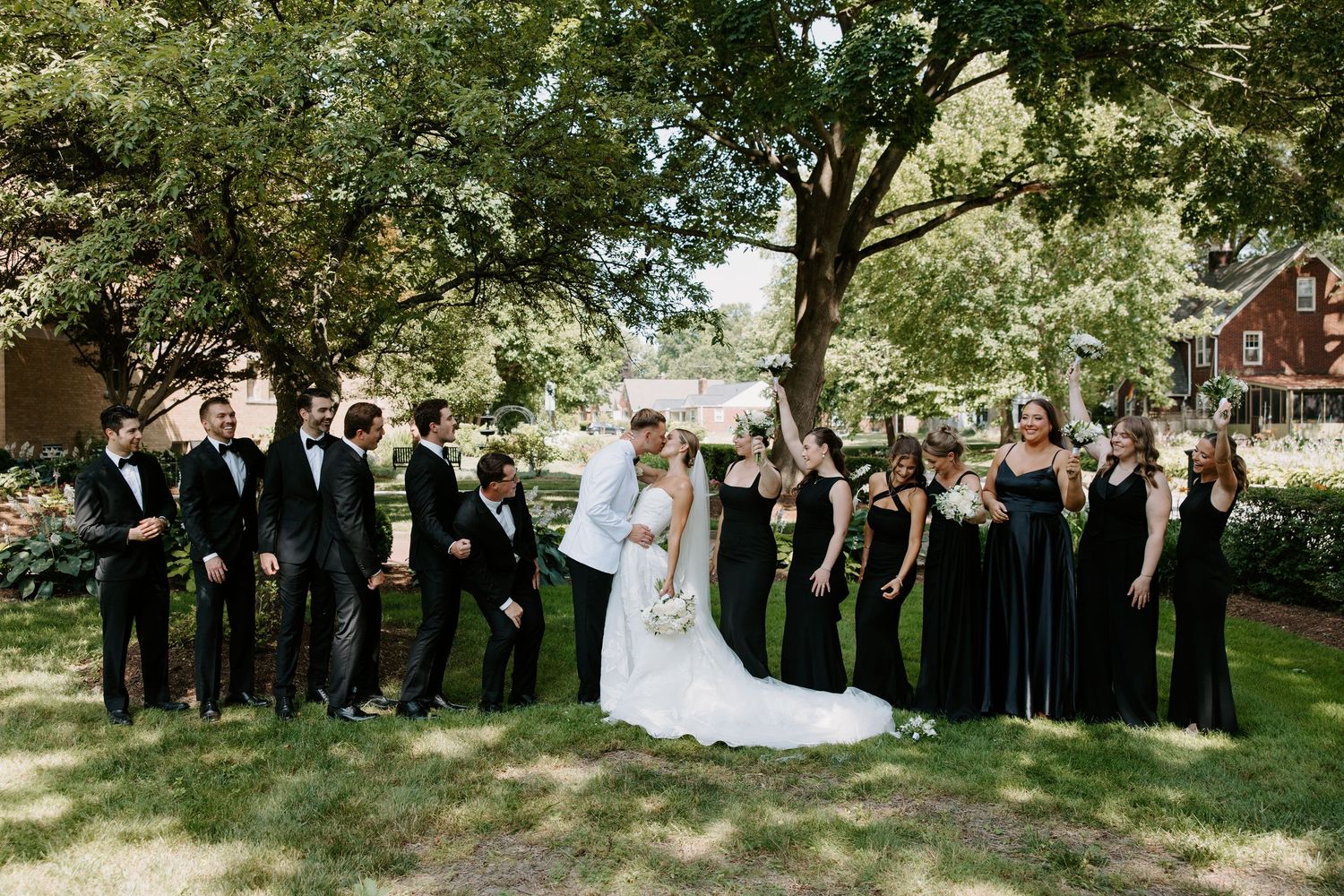 Wedding party in black attire poses with bride and groom under shady trees at outdoor venue.