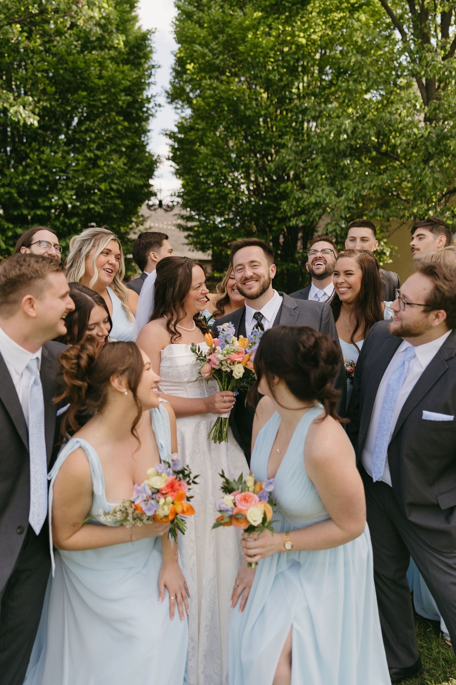 Bride and groom laugh with wedding party in light blue gowns and gray suits surrounded by colorful bouquets.