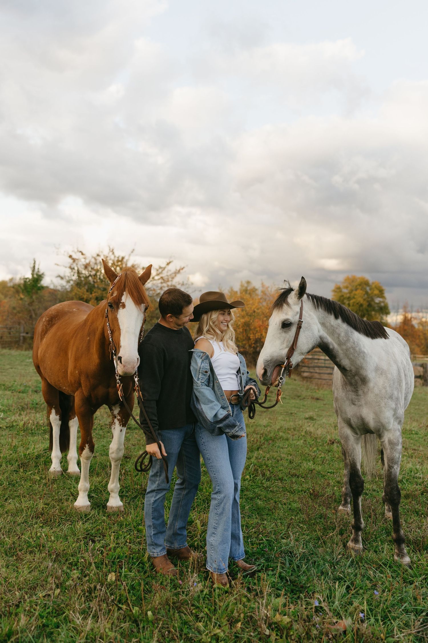 A couple stands with two horses, one brown and one gray, on a green field under a cloudy sky.