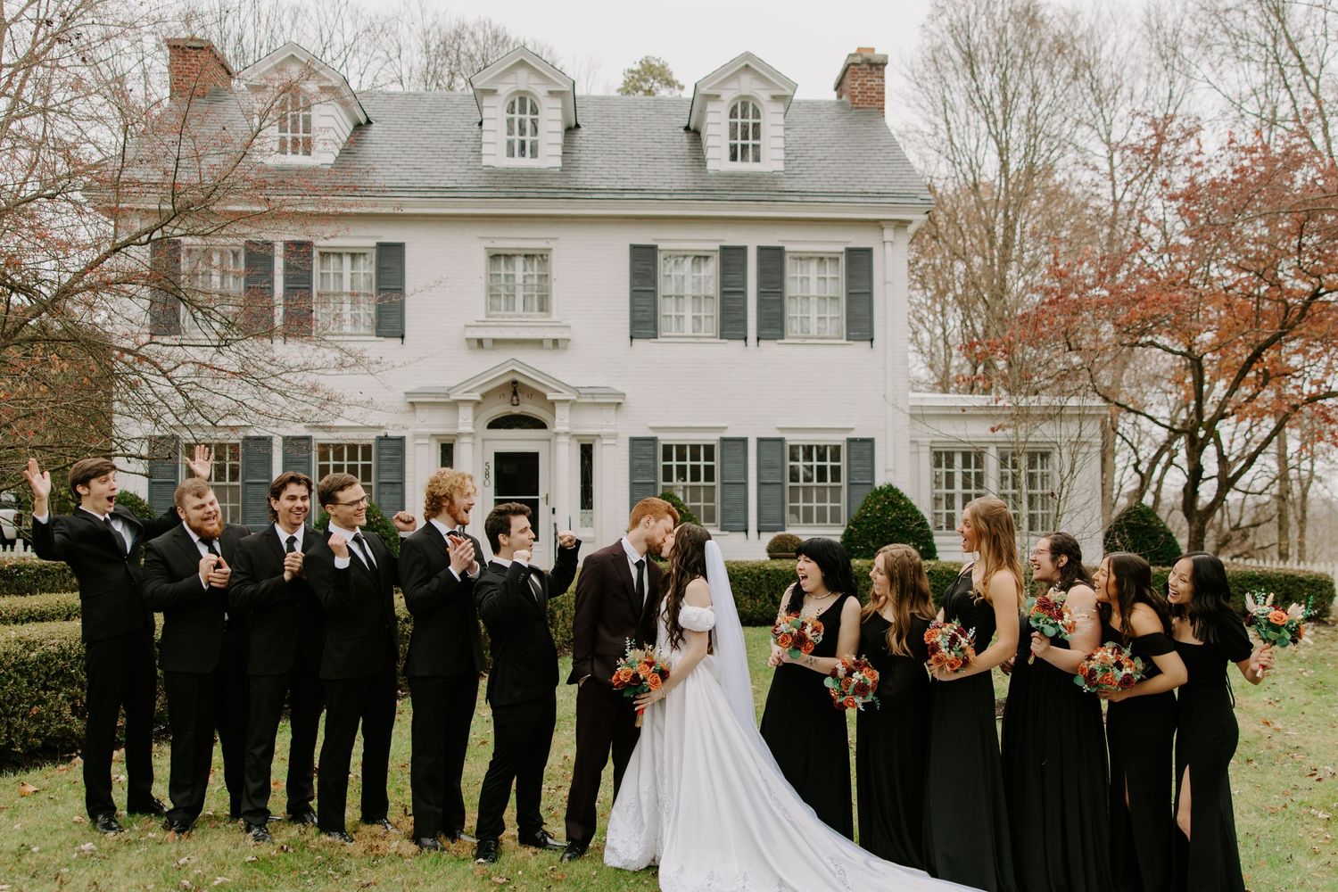 Wedding party in all-black attire celebrating with bride and groom in front of a white colonial manor house.