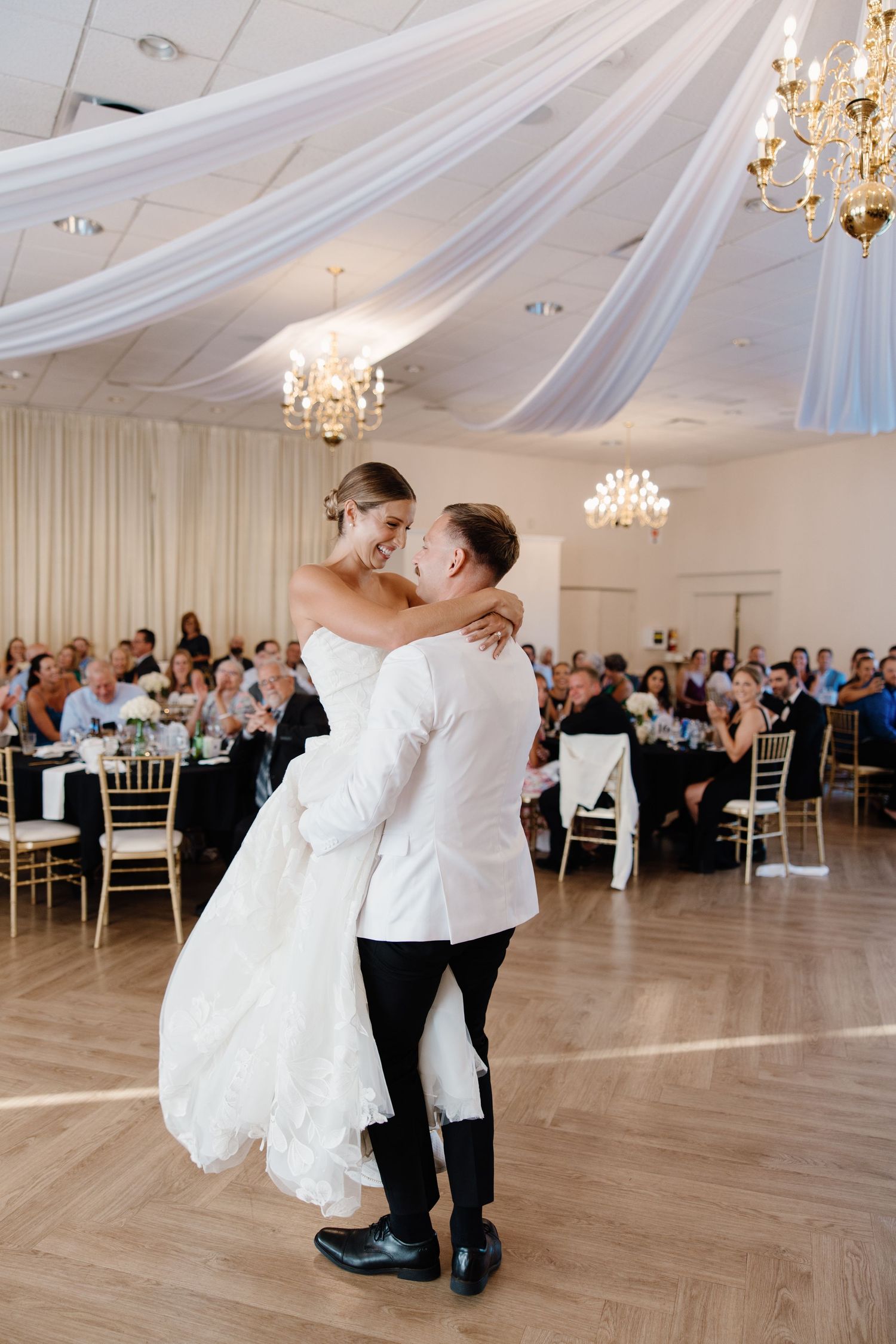 Groom lifts bride during their first dance in a beautifully draped ballroom filled with wedding guests.