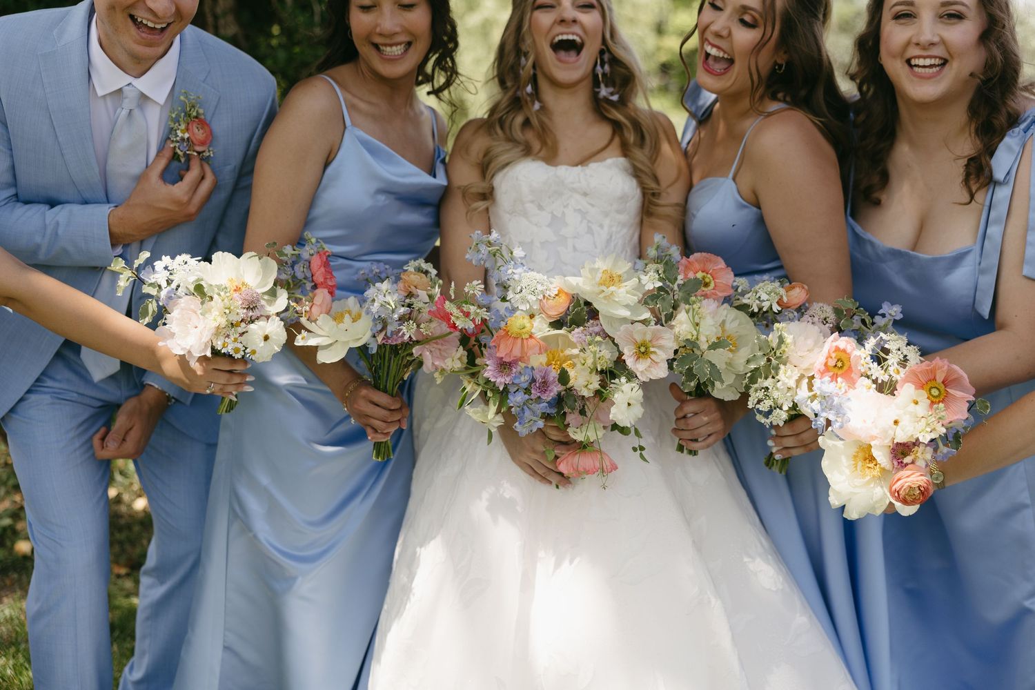 Bride in white gown laughing with bridesmaids in blue dresses holding colorful floral bouquets outdoors.