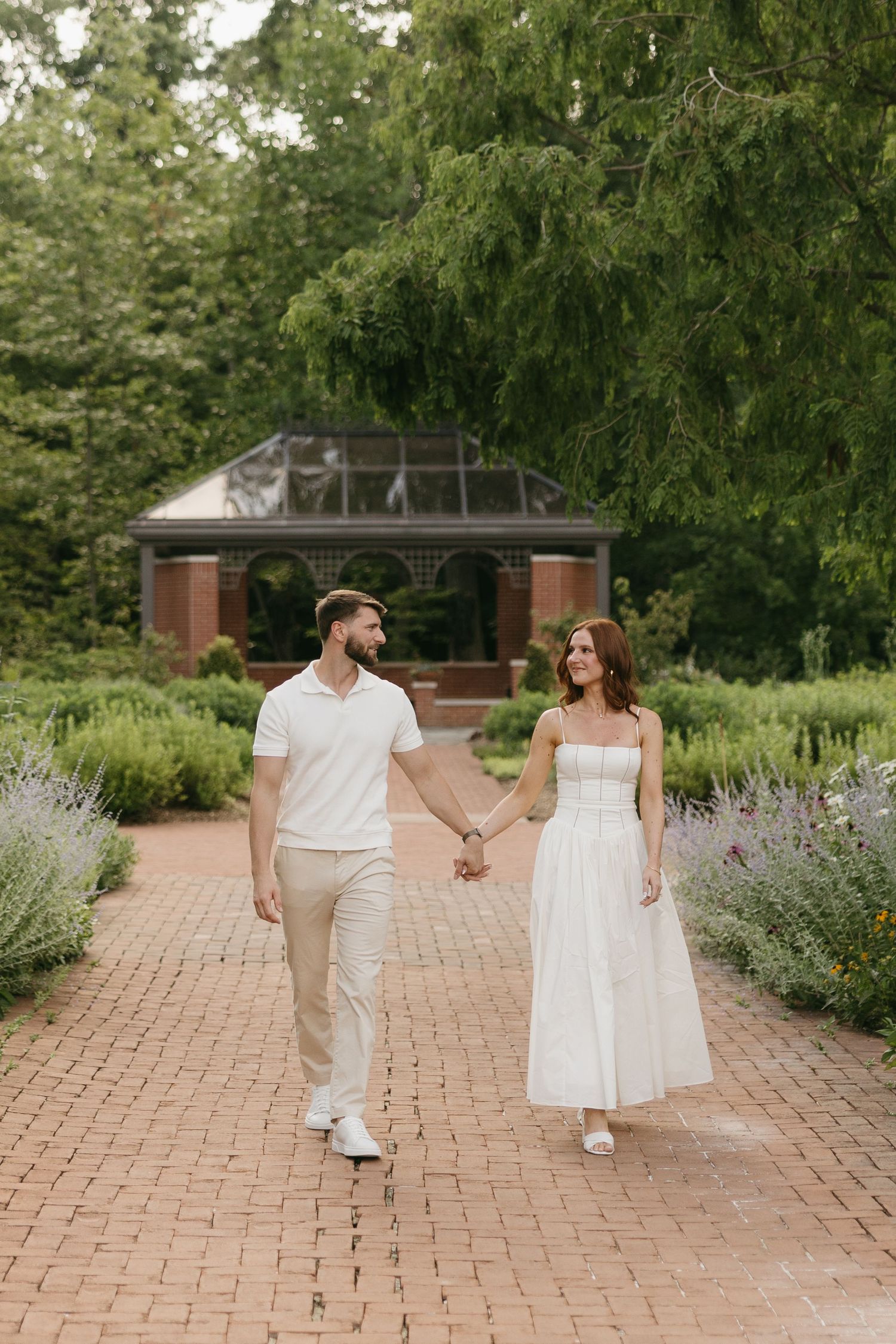 Couple in white outfits walk hand-in-hand on a brick path through a lush garden.