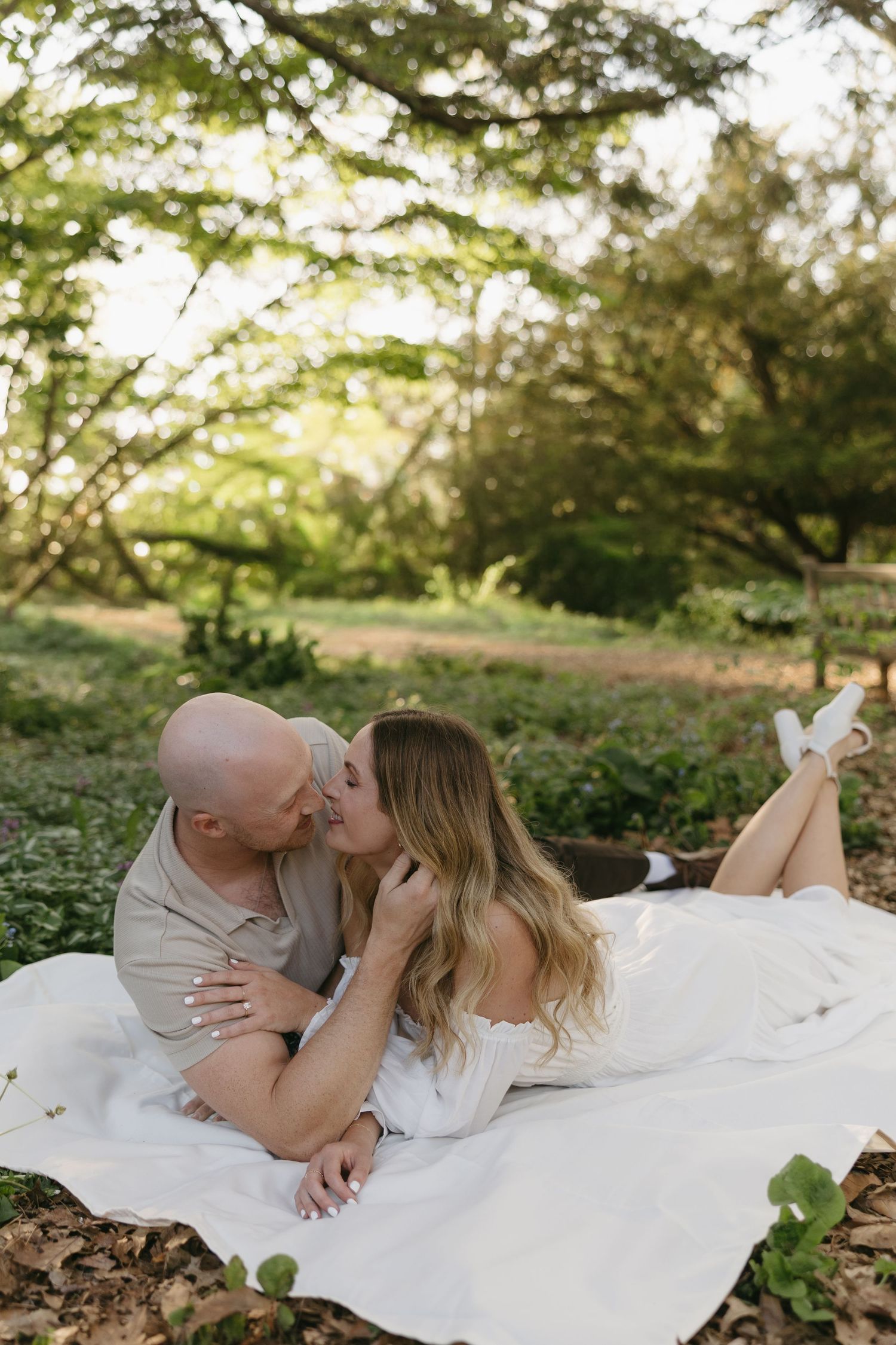Couple sharing a romantic kiss while lying on a white blanket in a sunlit garden setting.