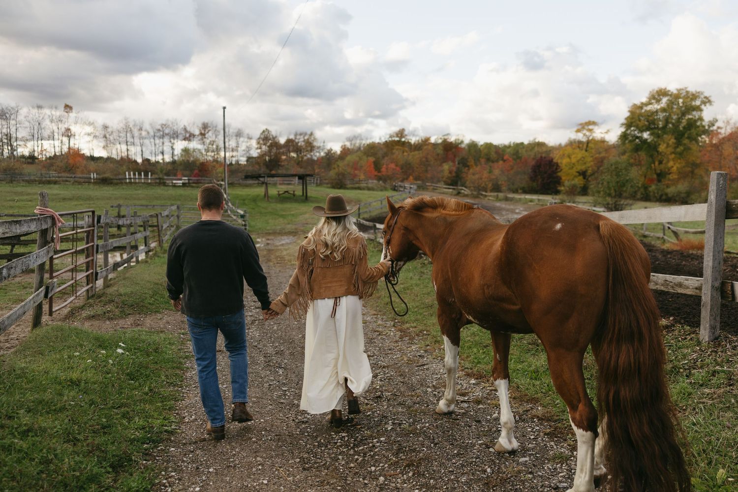 Couple walking hand in hand with a chestnut horse along a rural farm path surrounded by autumn foliage.