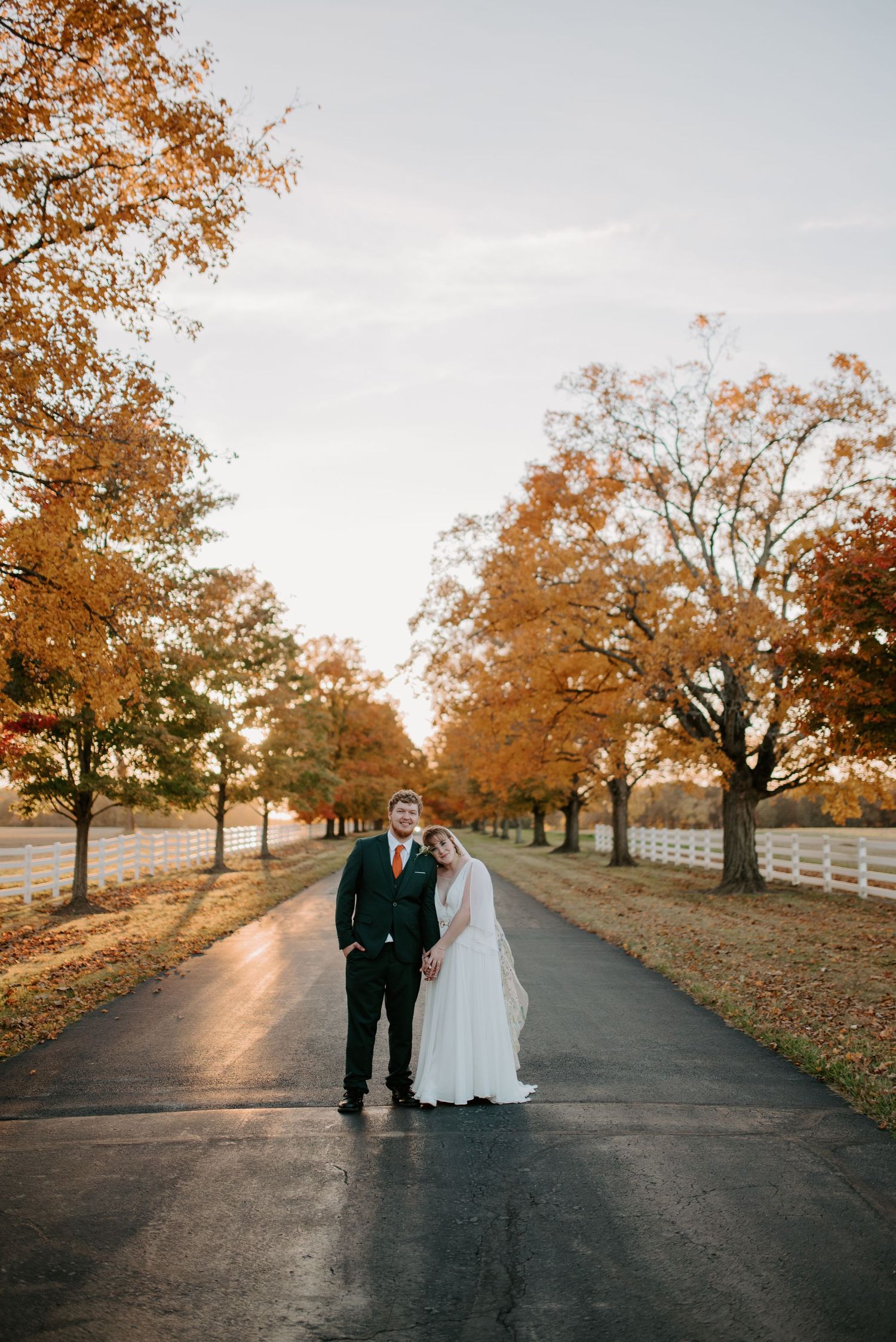 Bride and groom pose on a tree-lined autumn driveway with white fences and golden fall foliage.