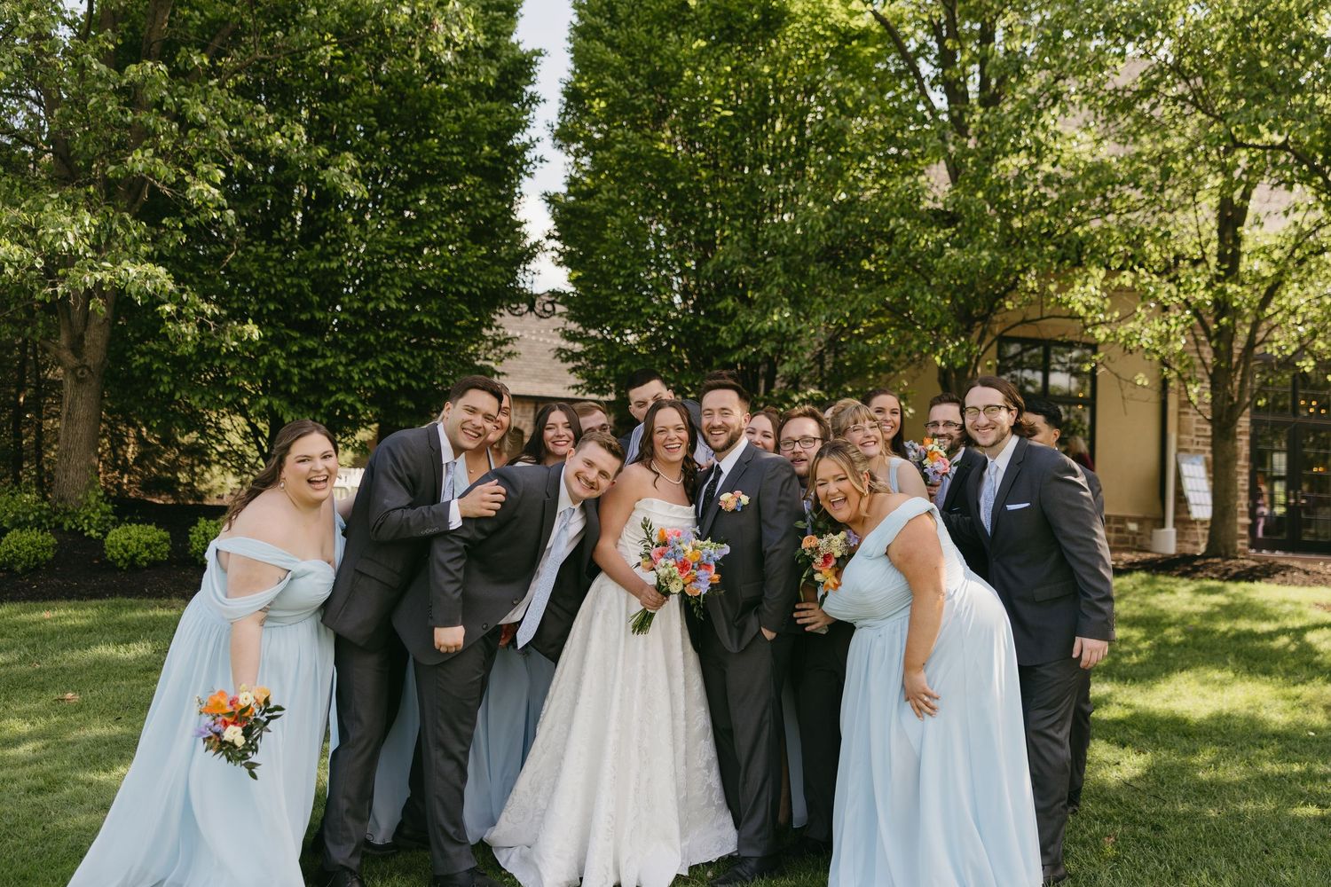 Joyful wedding party in blue bridesmaid dresses and gray suits pose together outdoors under lush green trees.