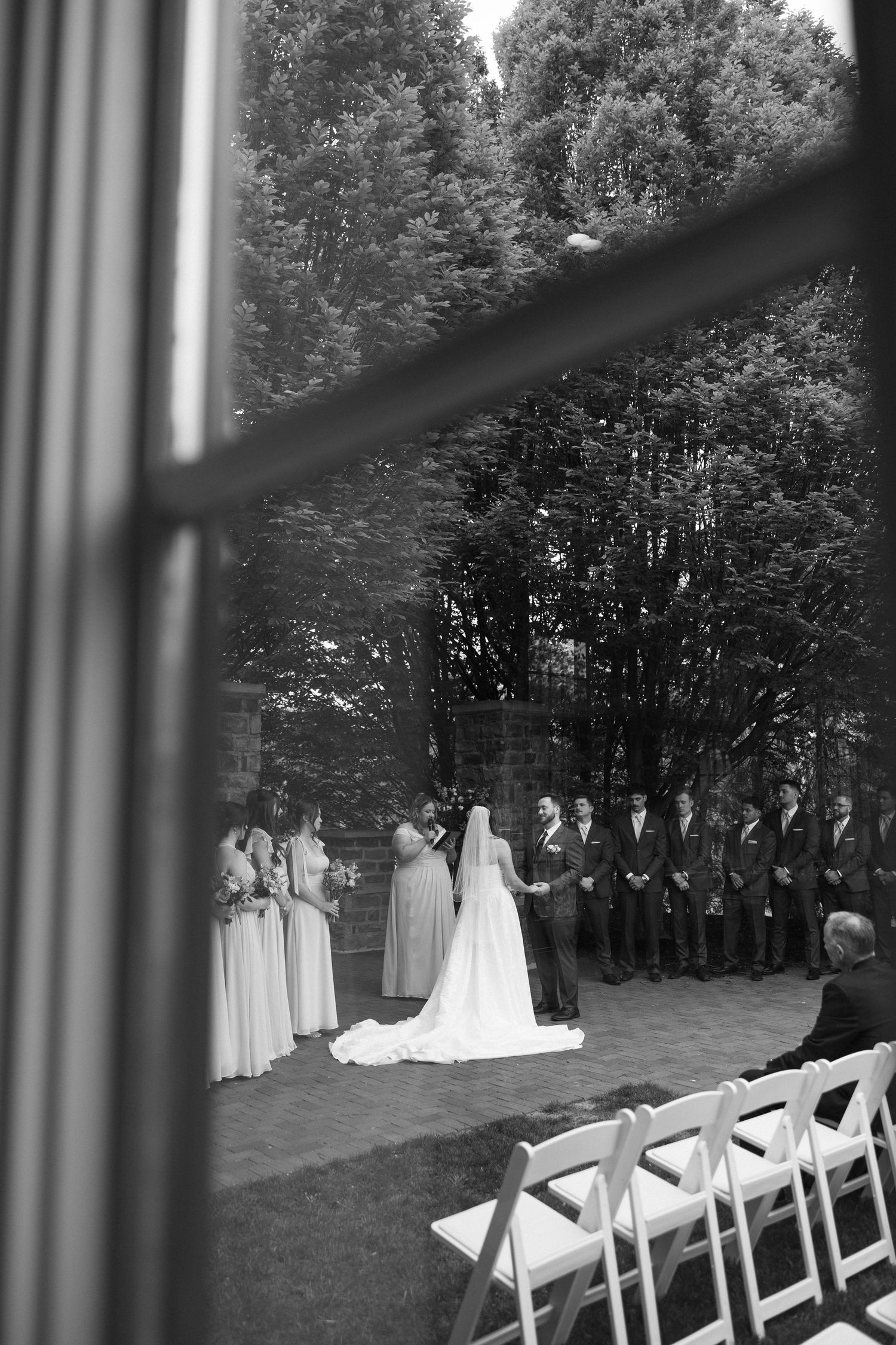 Black and white photo of wedding ceremony viewed through a window with bridal party and guests seated.