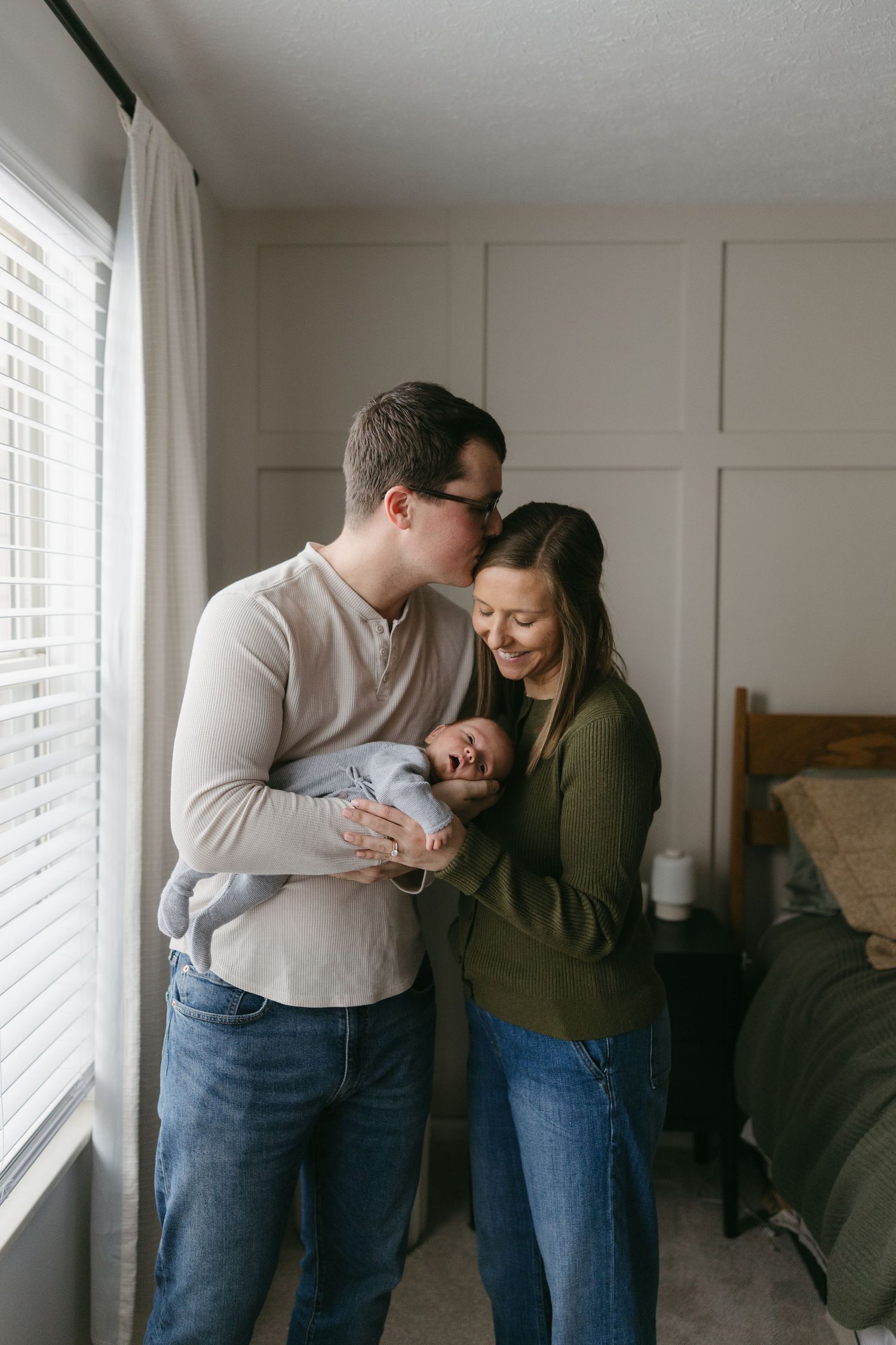 New parents standing by a window holding and kissing their crying newborn baby.