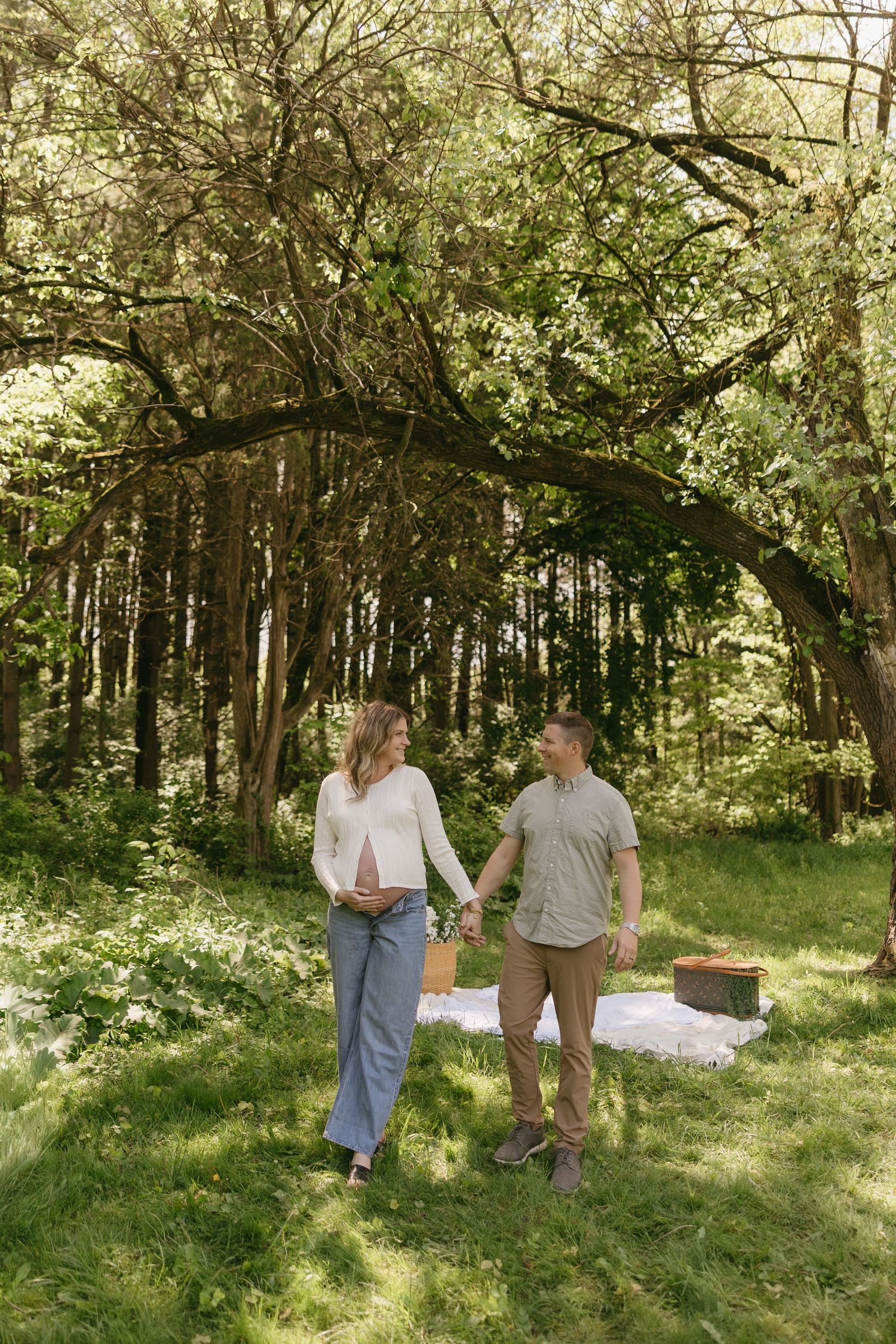 Pregnant couple holding hands and walking together in a sunlit green forest.