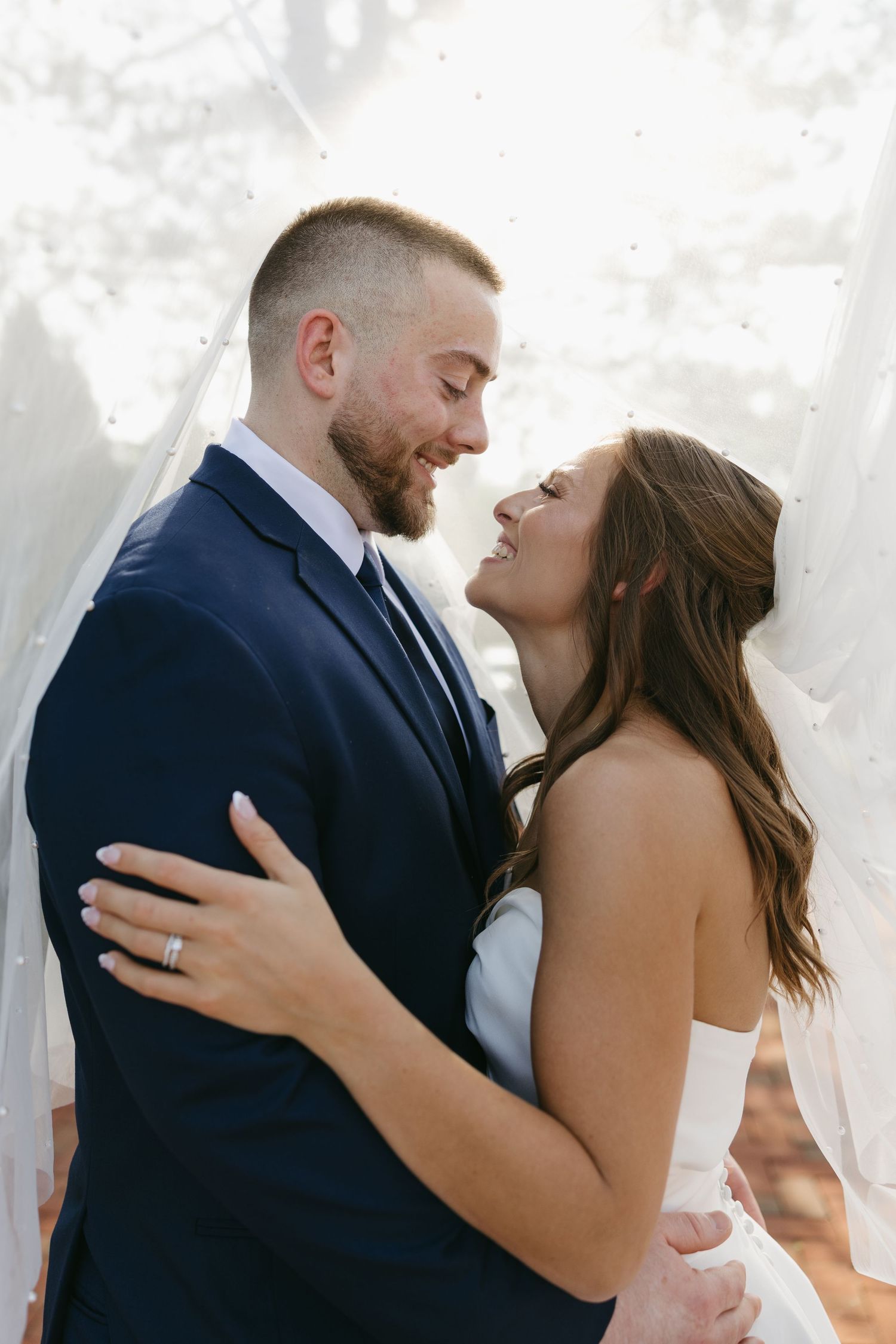 Bride and groom share romantic moment under sheer veil, nearly kissing in soft natural outdoor light.