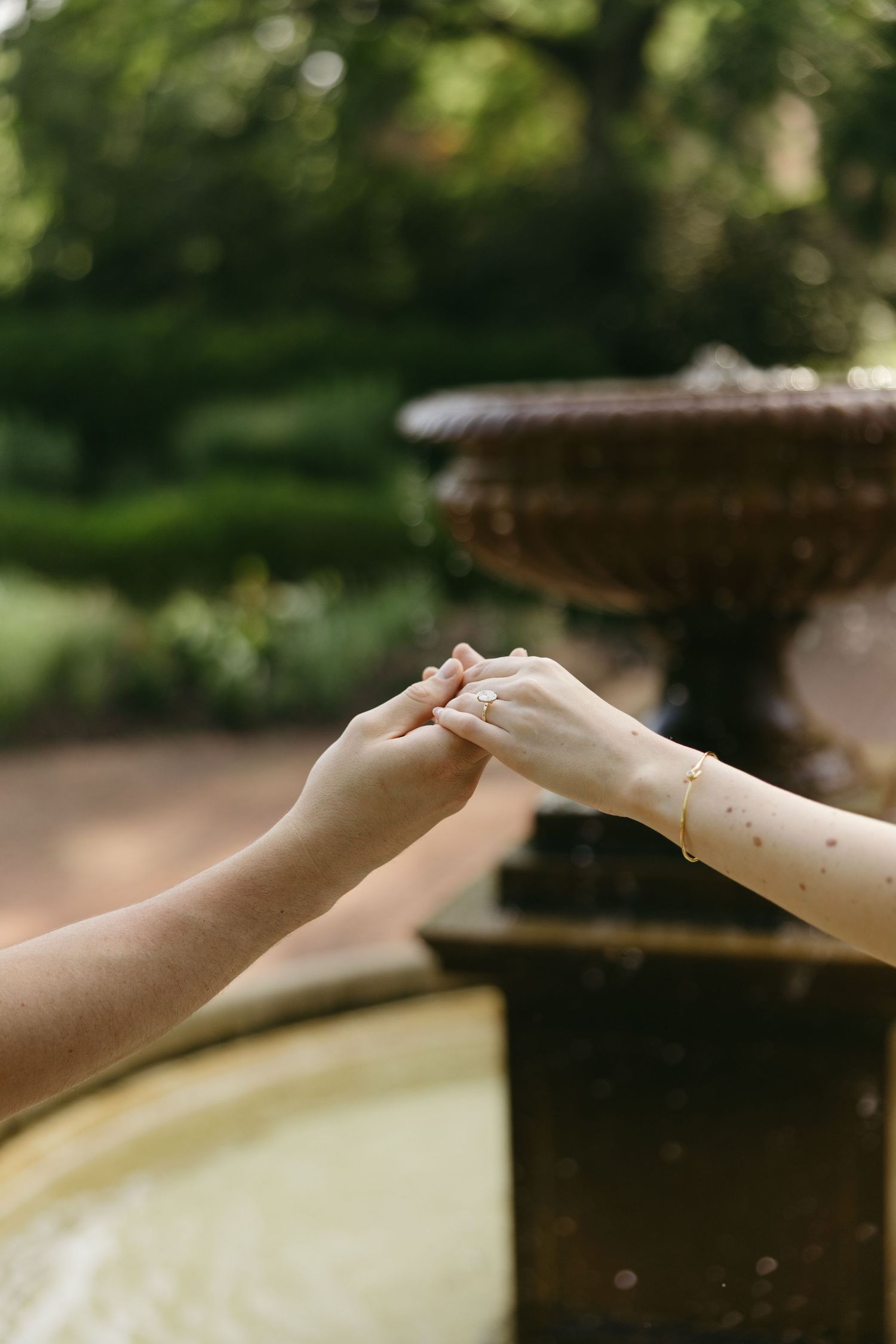 Close-up of engaged couple holding hands showing diamond ring near a stone garden fountain.