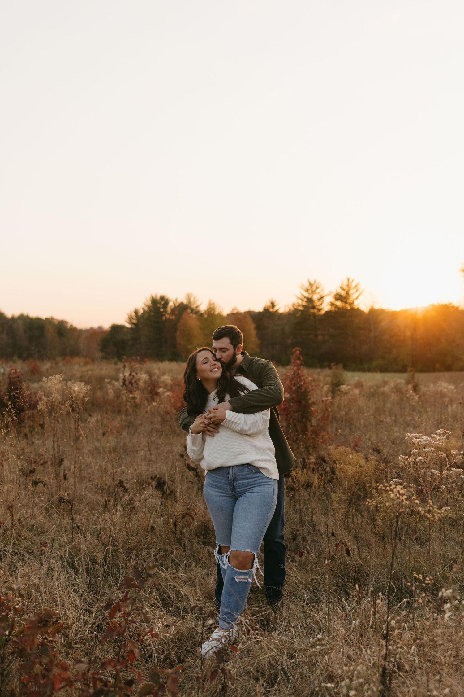 Man embracing woman from behind in a golden sunset field, both smiling in casual fall outfits.